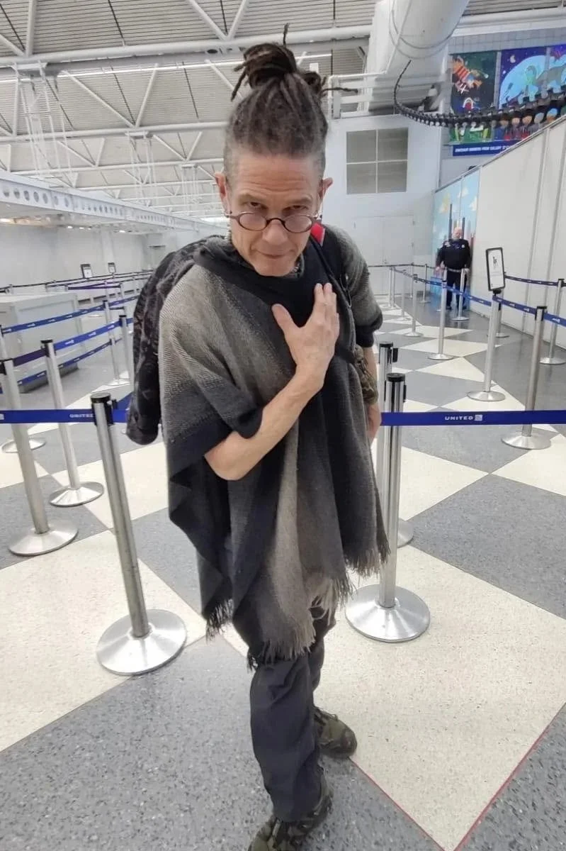 Taylor, wearing purple glasses, dressed in dark clothing, with dreadlocks, standing in an airport terminal. He is holding his chest with one hand and looking at the camera with a serious expression.