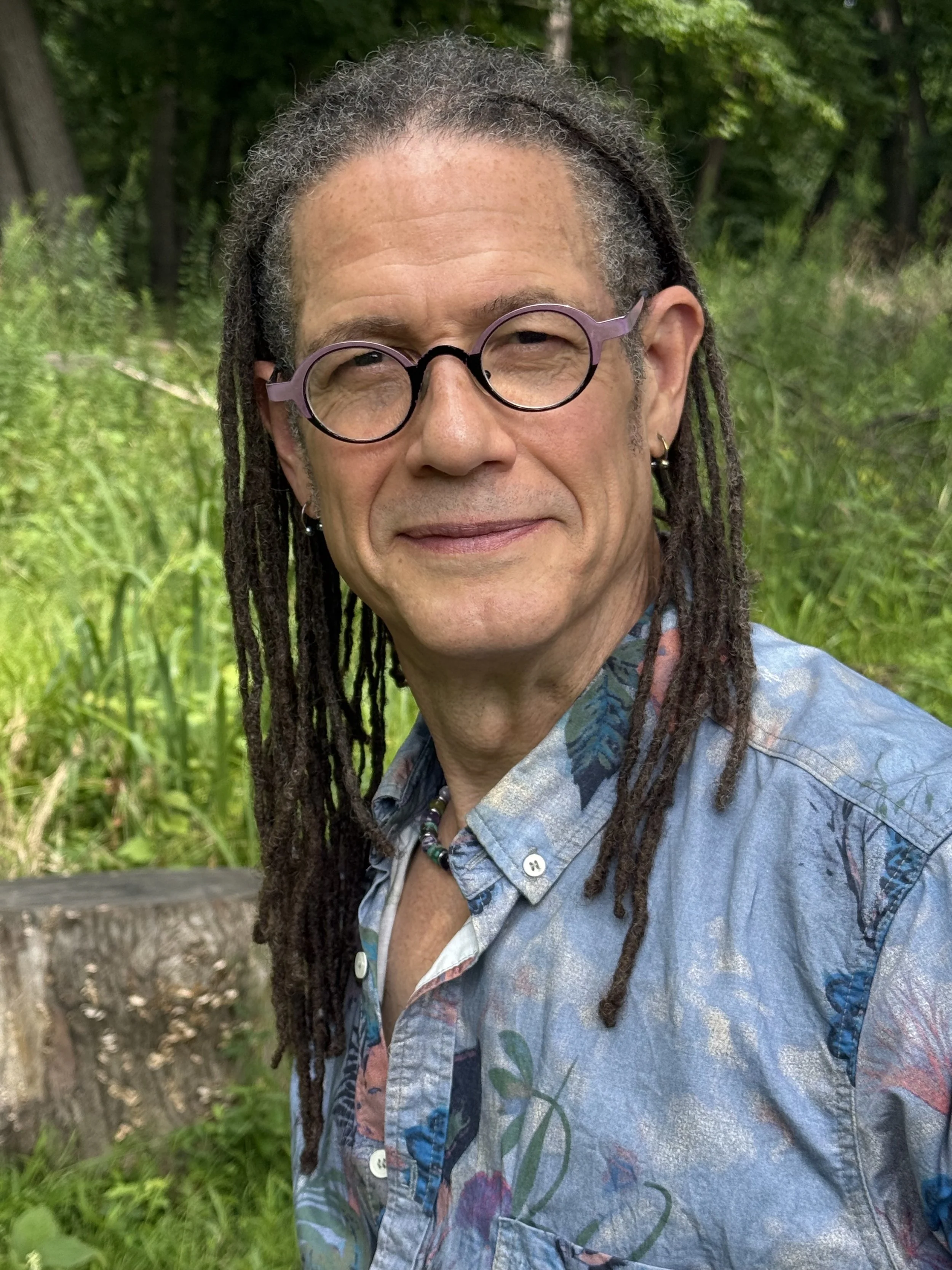 meet M. Taylor, Life Coach. In this photo of him, he sports his recognizable dreadlocks, glasses, and a colorful button-up shirt, standing outside and smiling at you in front of a forest of green trees in the background.