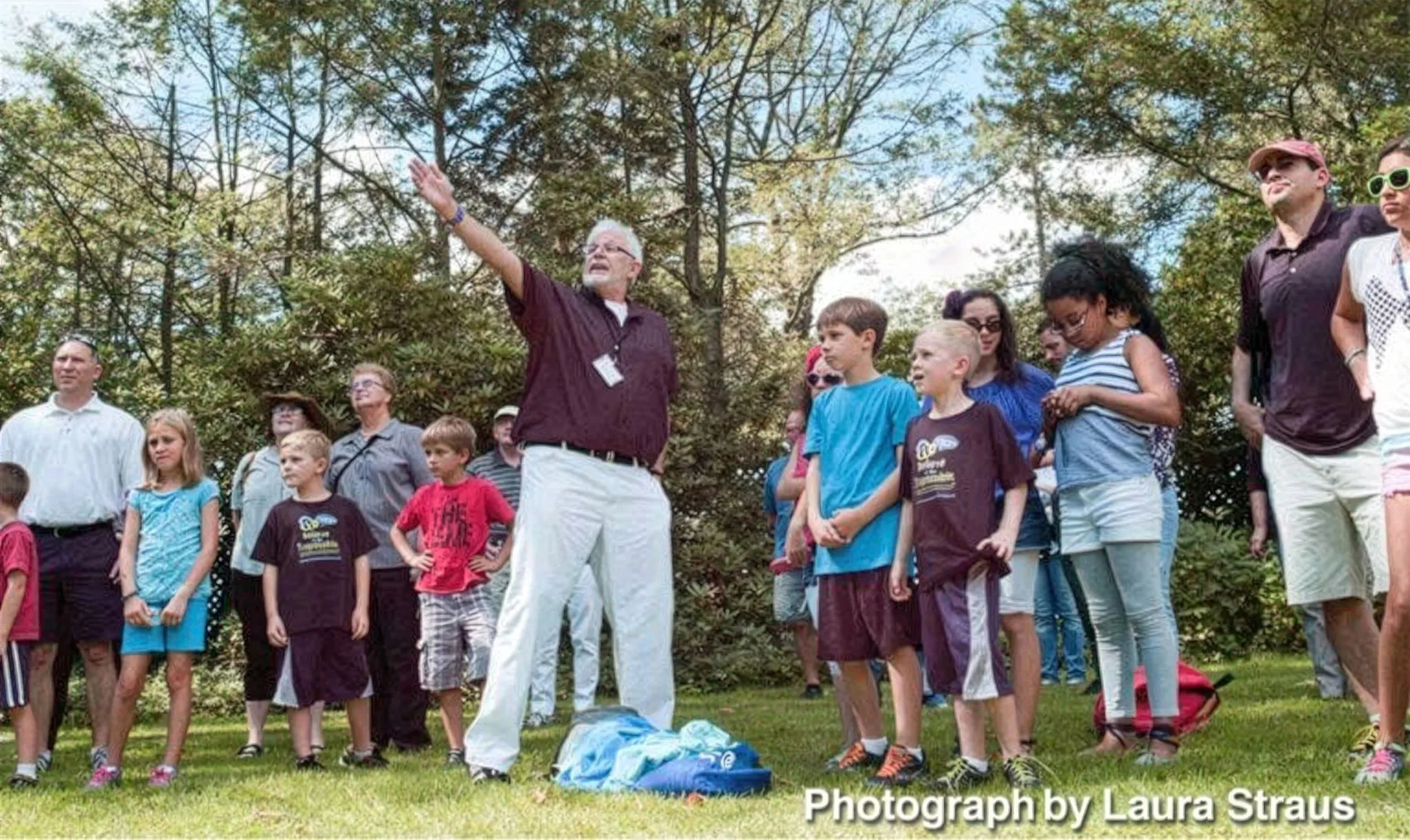 A man with glasses, white hair, and a beard is speaking or giving instructions to a group of children and adults outdoors in a park or garden with trees in the background.