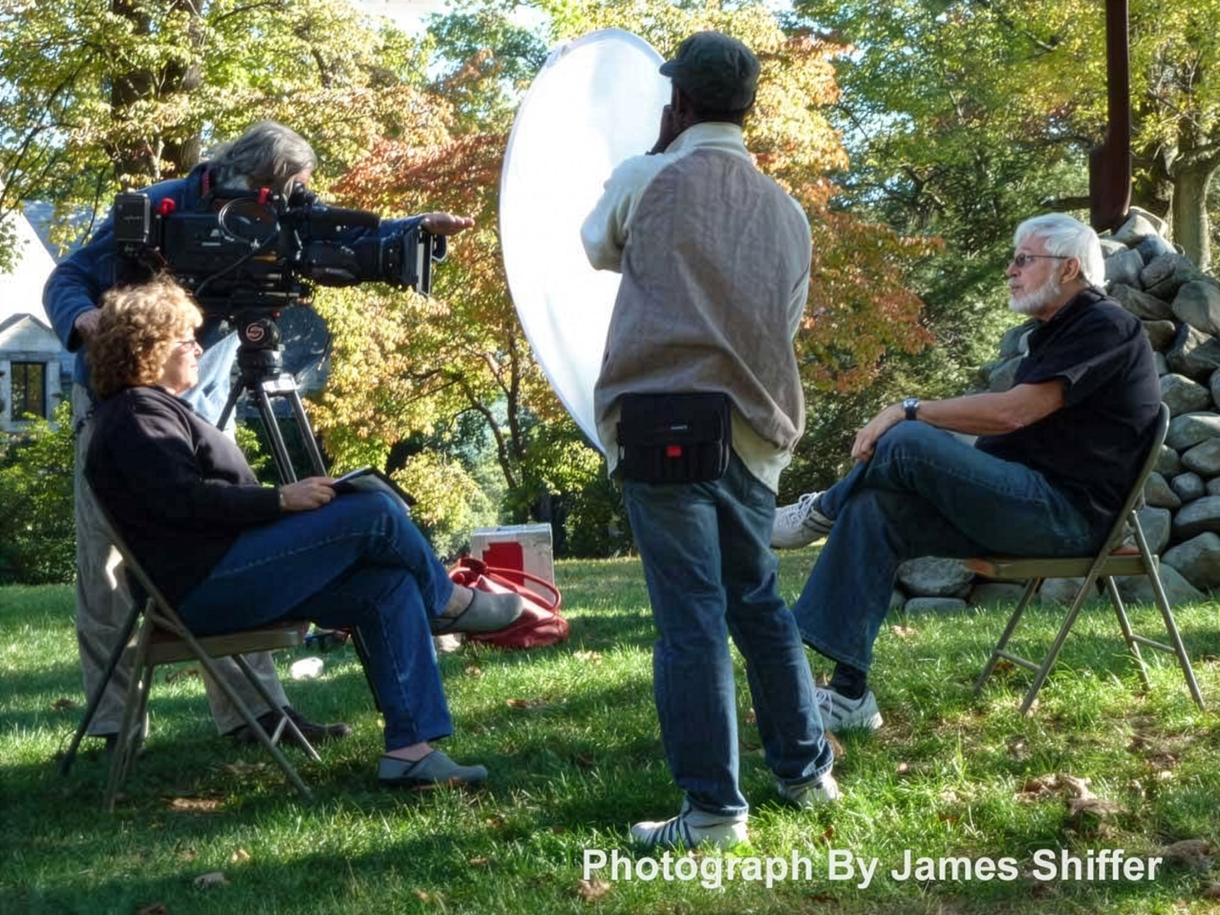 A group of five people engaged in a photography shoot outdoors during autumn. One woman with headphones operates a camera on a tripod, while a seated woman and a seated man are being photographed against a backdrop of colorful fall trees. The photographer and the seated individuals are surrounded by green grass and fallen leaves.