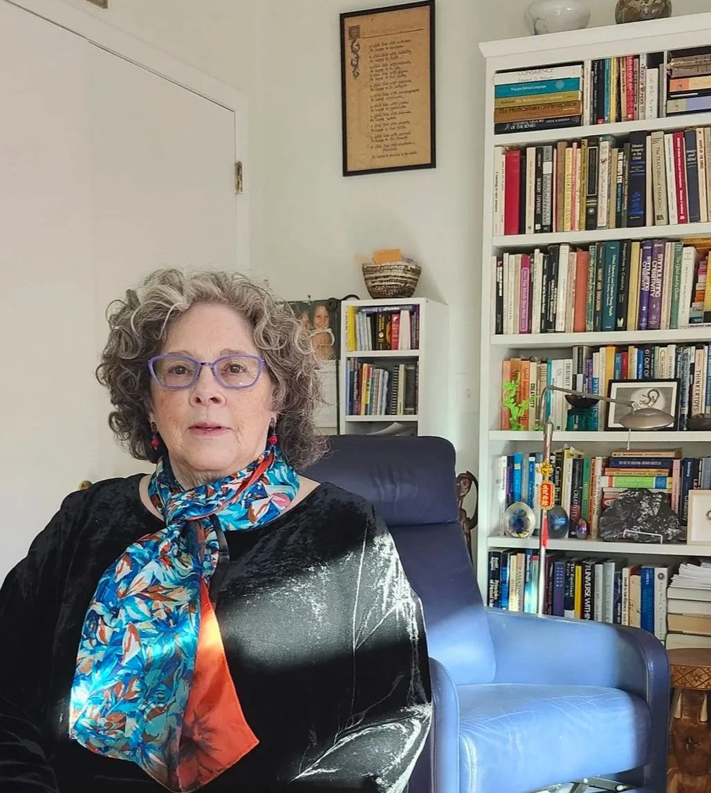 A woman with curly gray hair and purple glasses sitting in a room filled with bookshelves, a blue armchair, and artwork on the wall.