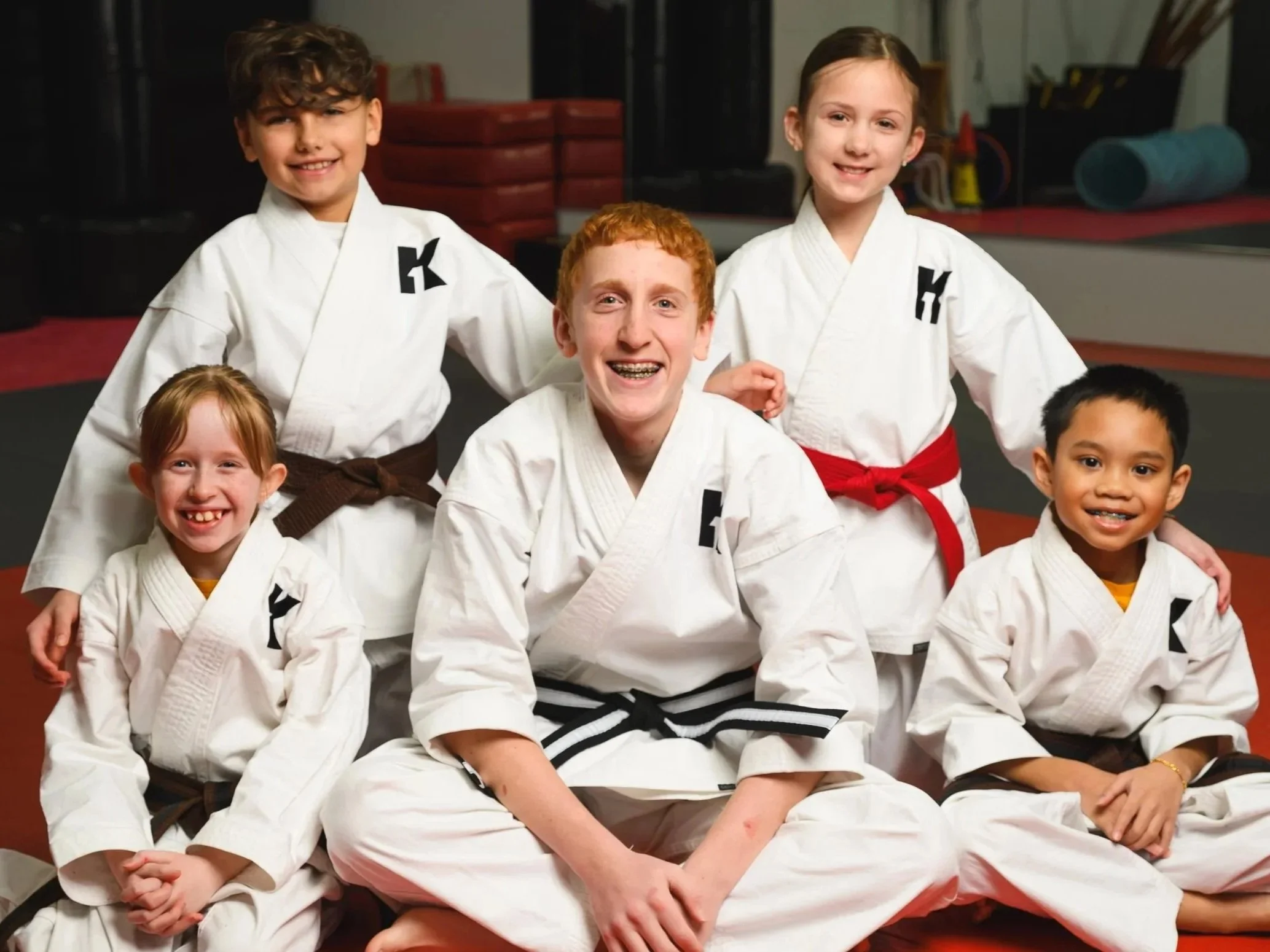 Group of children in martial arts uniforms smiling, sitting on a gym floor in a dojo.