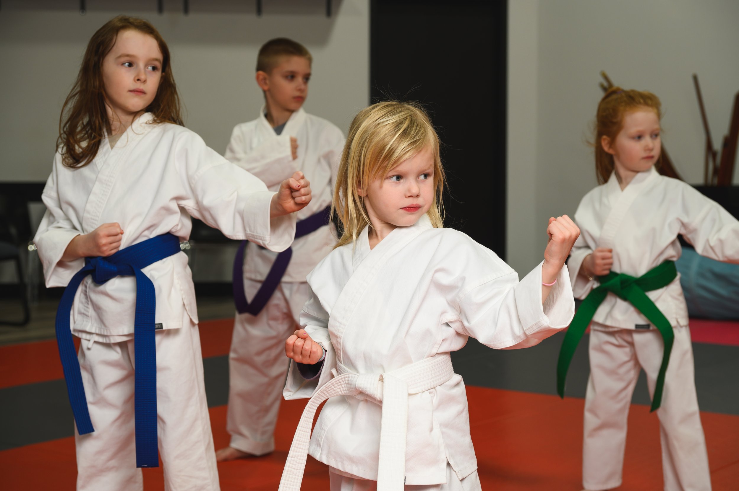 Young children practicing martial arts in white uniforms, some with colored belts, in a dojo or gym setting.