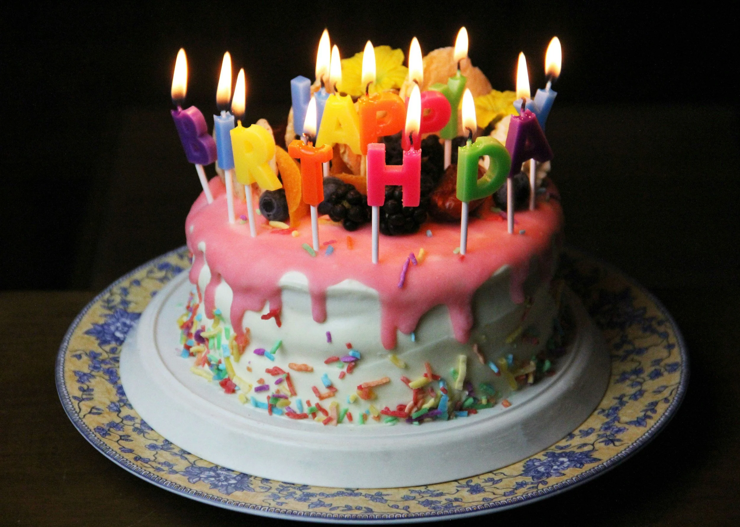 A birthday cake with pink icing, colorful sprinkles, and lit multicolored candles spelling out 'HAPPY BIRTHDAY'.