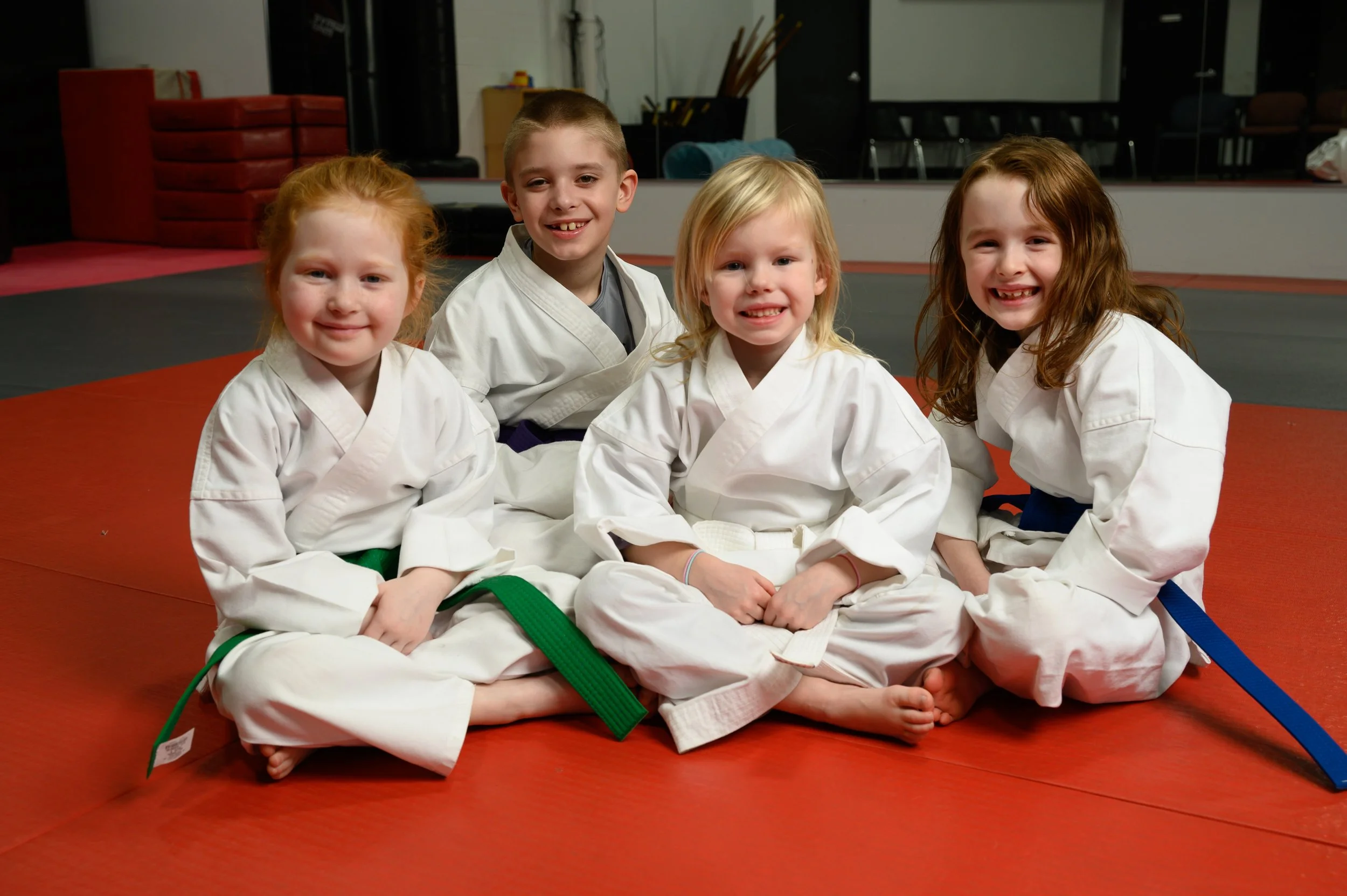 Five children in white karate uniforms sitting on red and black mats inside a martial arts dojo, smiling at the camera.