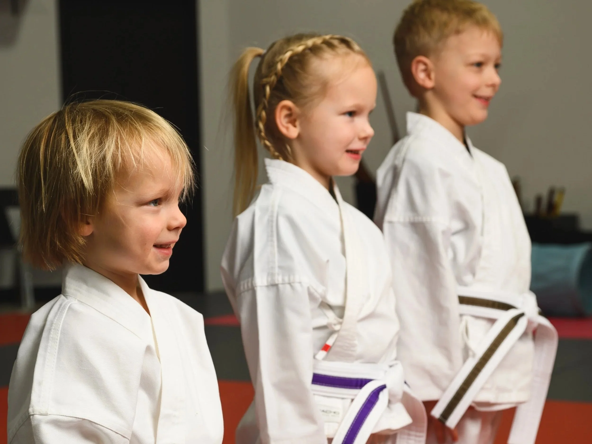 Four children wearing white martial arts uniforms standing in line, holding wooden sticks in a martial arts class or practice session.