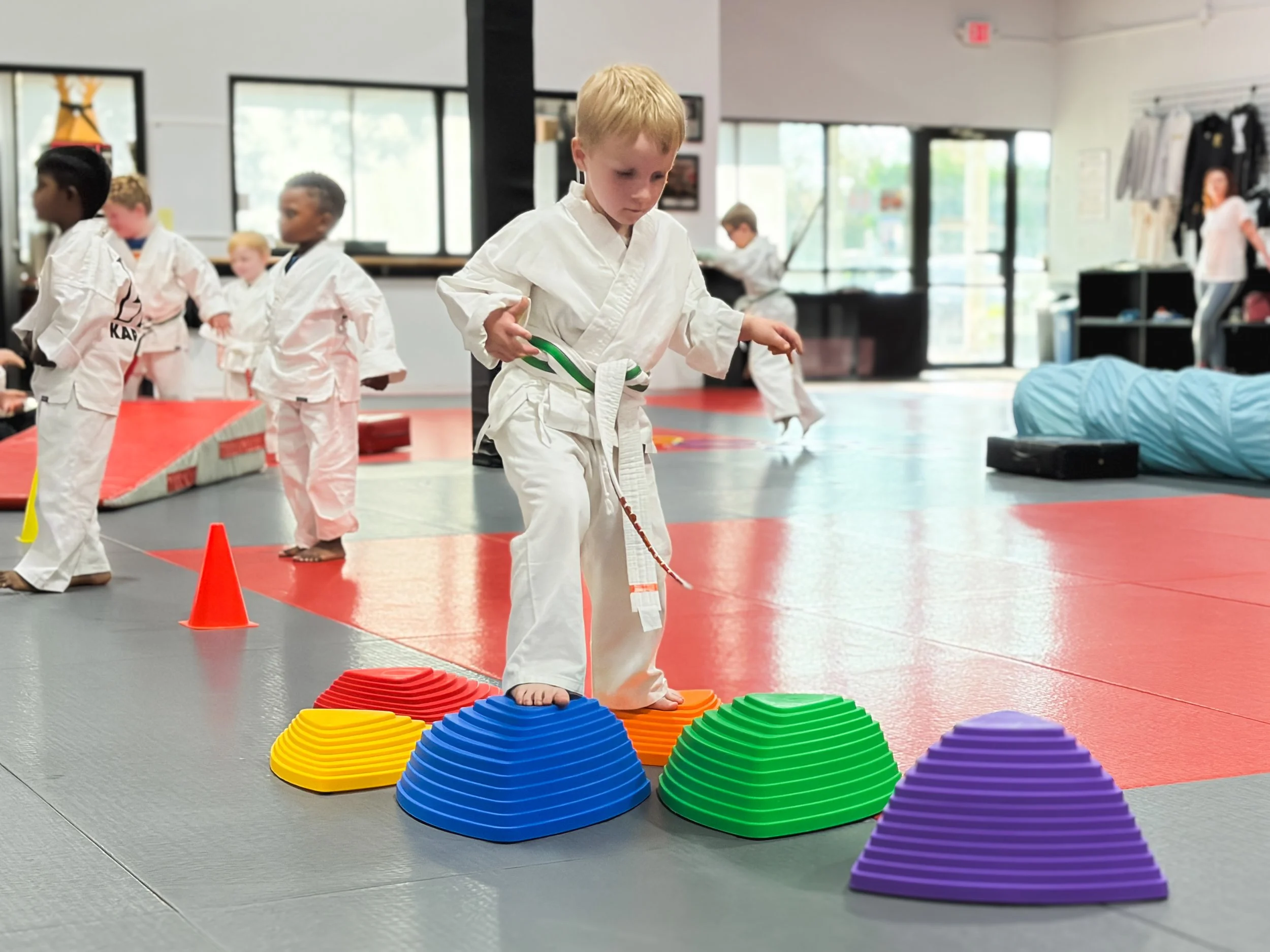 Young boy in a white martial arts uniform with a green belt balancing on colorful plastic cones in a martial arts studio. Several children in martial arts uniforms are in the background, with one adult woman standing near the wall.