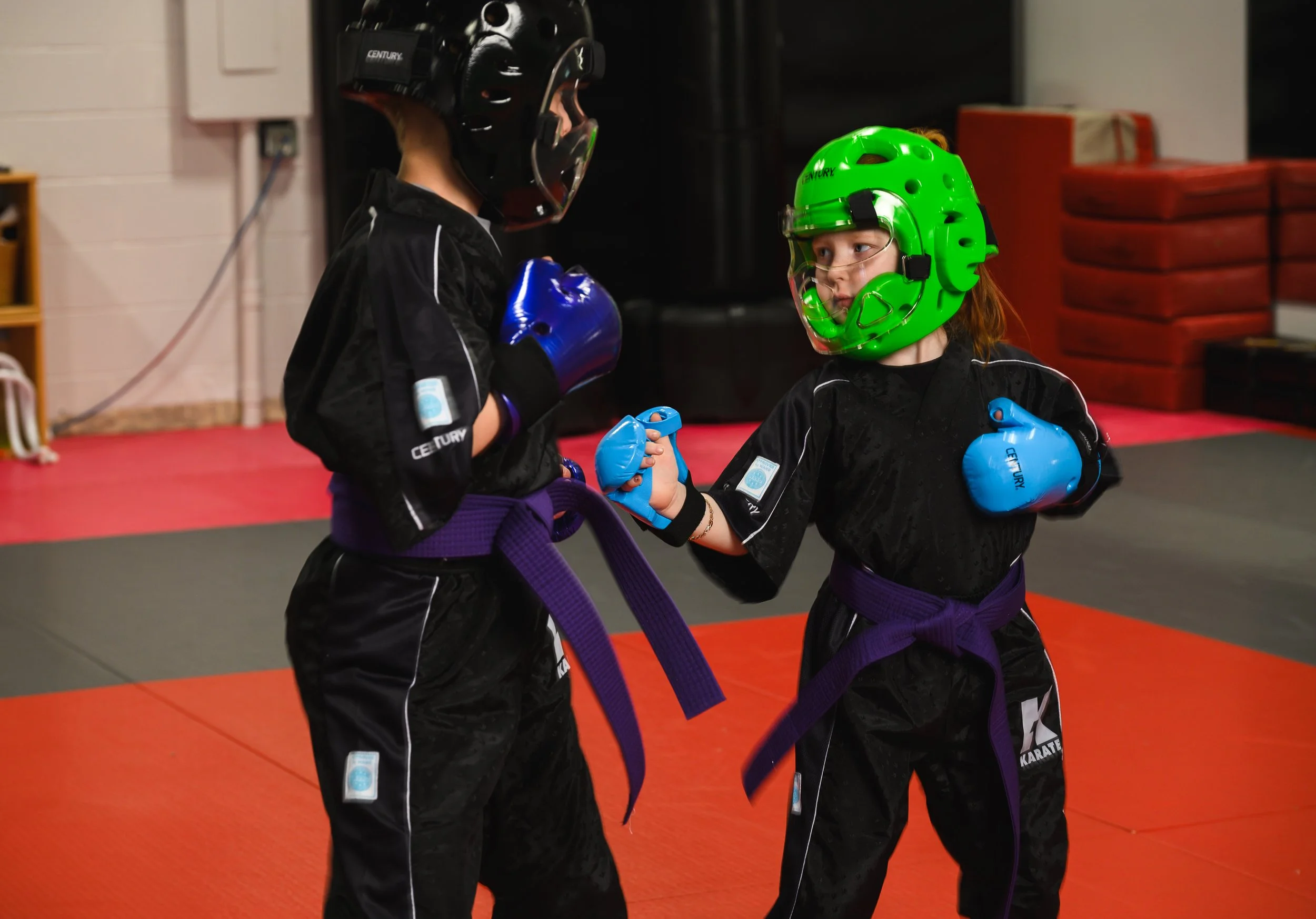 Two children wearing protective gear, including helmets and gloves, practicing martial arts in a gym with red mats.