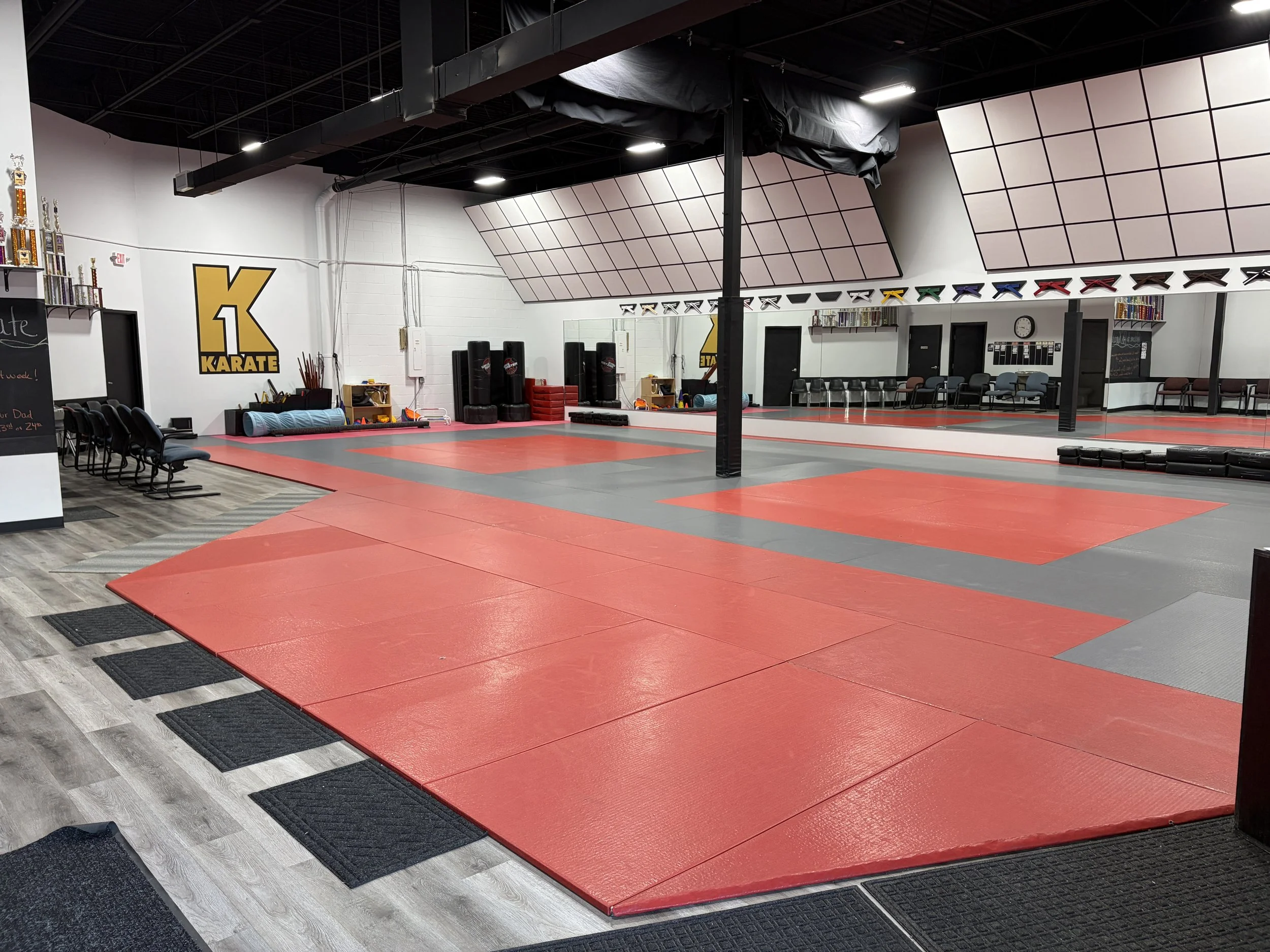 An indoor martial arts or karate training area with red and gray mats, black punching bags, trophies on a shelf, and colored belts hanging on display. There are black chairs in the background along the wall, and the wall has a large yellow and black 'K Karate' sign.