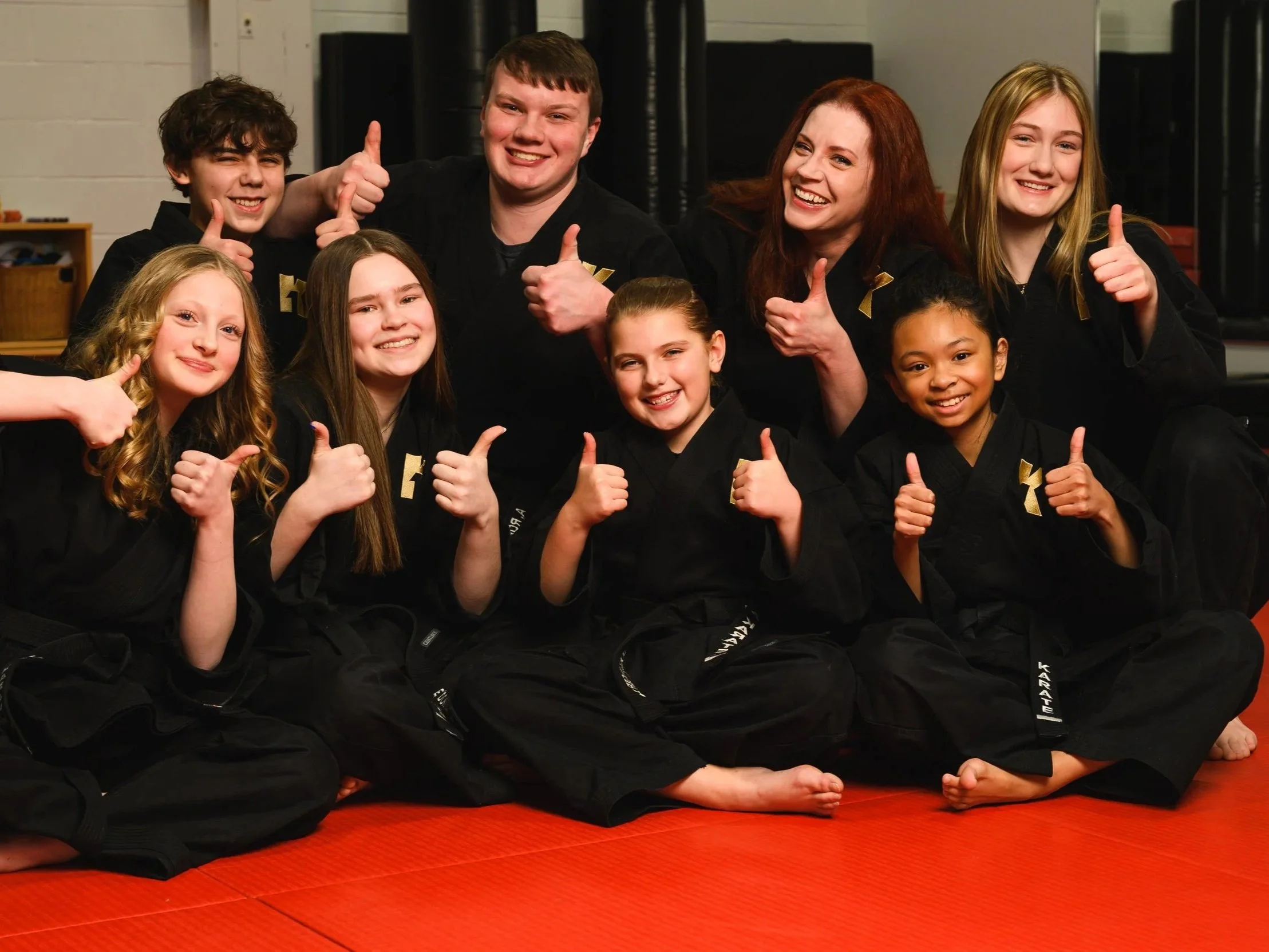 Group of children and an adult in martial arts uniforms sitting on a red mat, smiling and giving thumbs up.