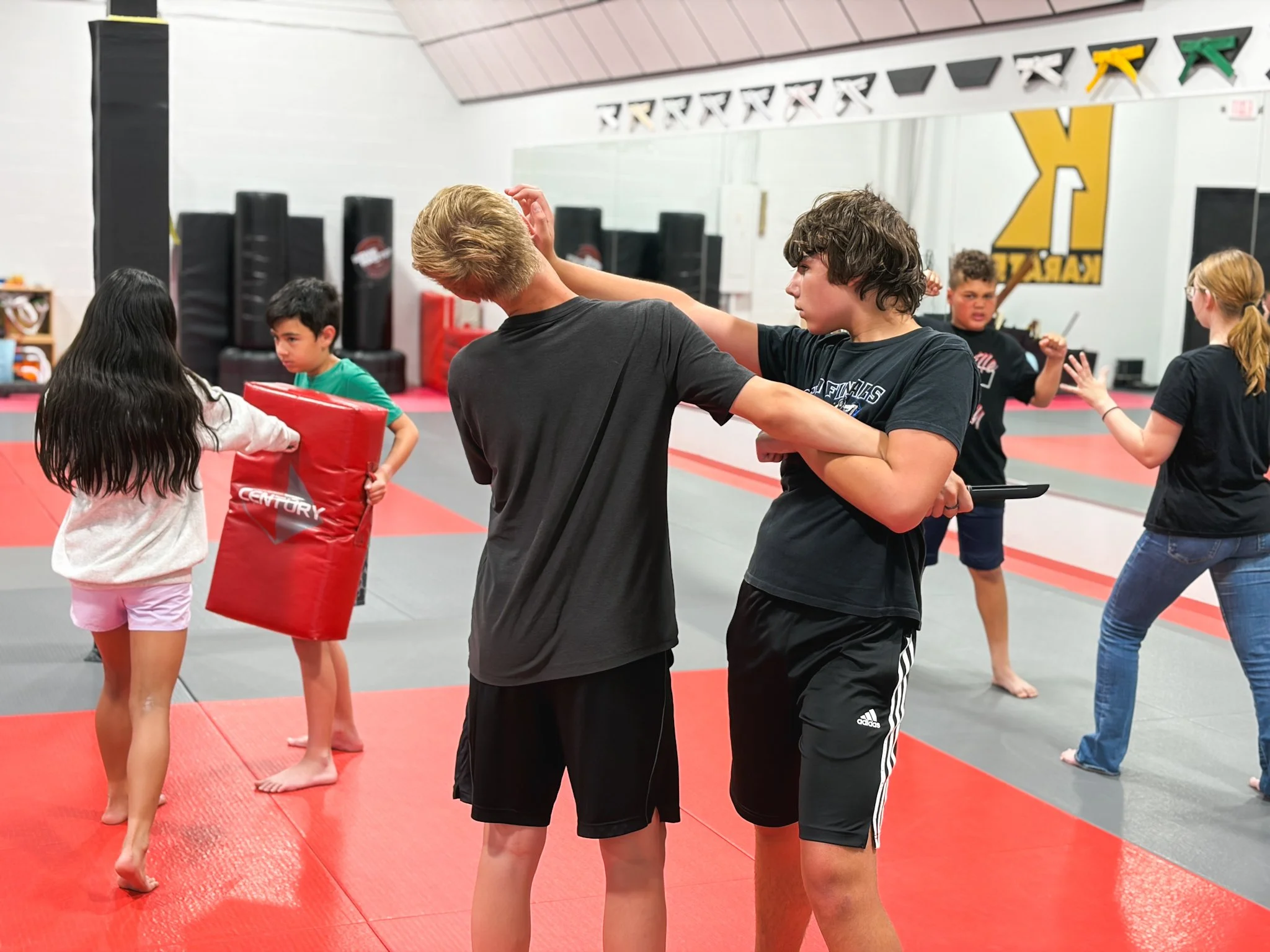 A group of children and a coach practicing martial arts in a gym, with some children holding pads and others sparring.