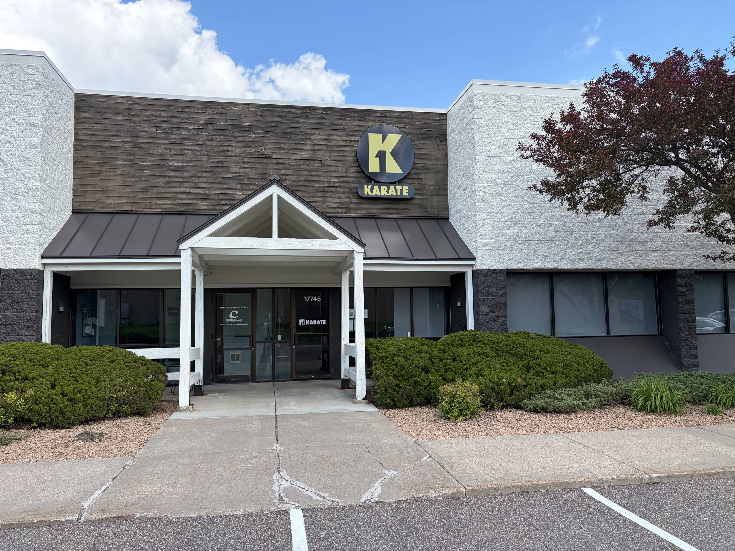 Front of a commercial building with a sign that reads 'K1 Karate' on the upper part of the facade, a small porch with a gable roof, surrounded by well-maintained bushes and a parking lot in front, against a blue sky with some clouds.