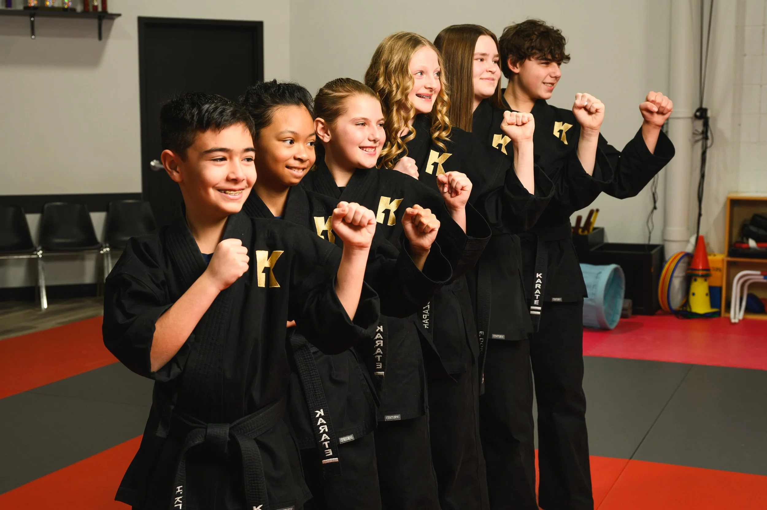 Group of six children in karate uniforms standing side by side, smiling and making fists, in a martial arts dojo with black belts and various training equipment in the background.