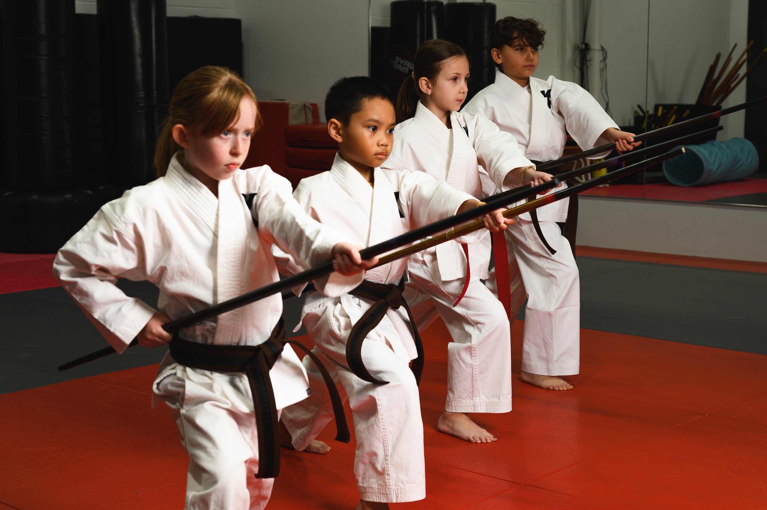 Five young martial arts students in white uniforms with black belts practicing sword techniques in a dojo, standing with swords extended forward on a red floor mat.