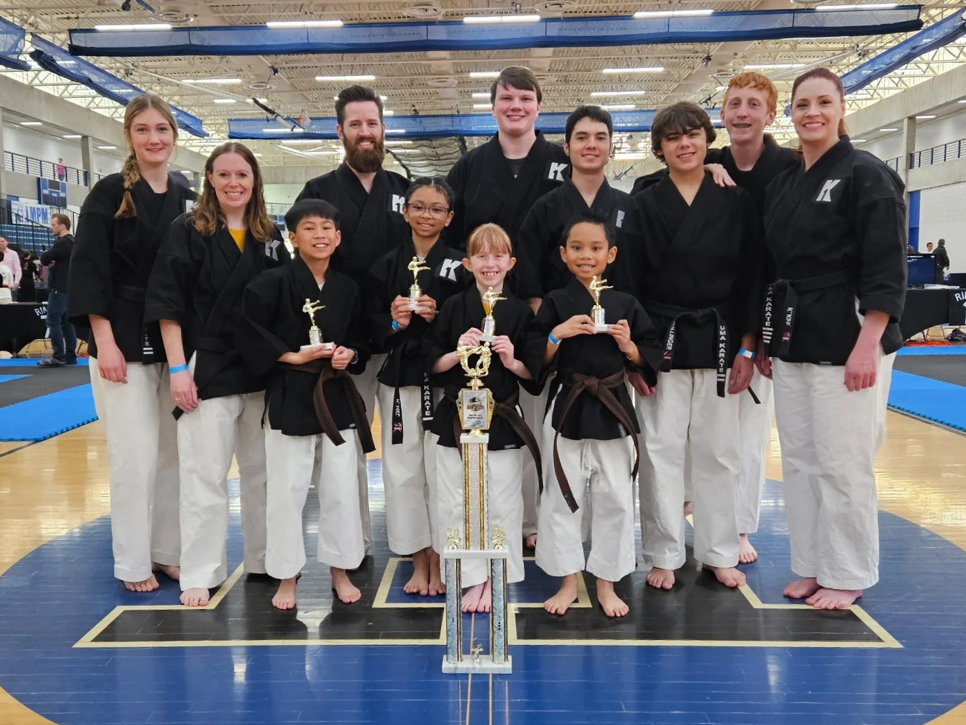 A group of martial artists, both adults and children, wearing black uniforms with brown belts standing together in a gymnasium. The children are holding trophies and a large trophy is placed on the ground in front. The group is smiling, showing they have won a competition.