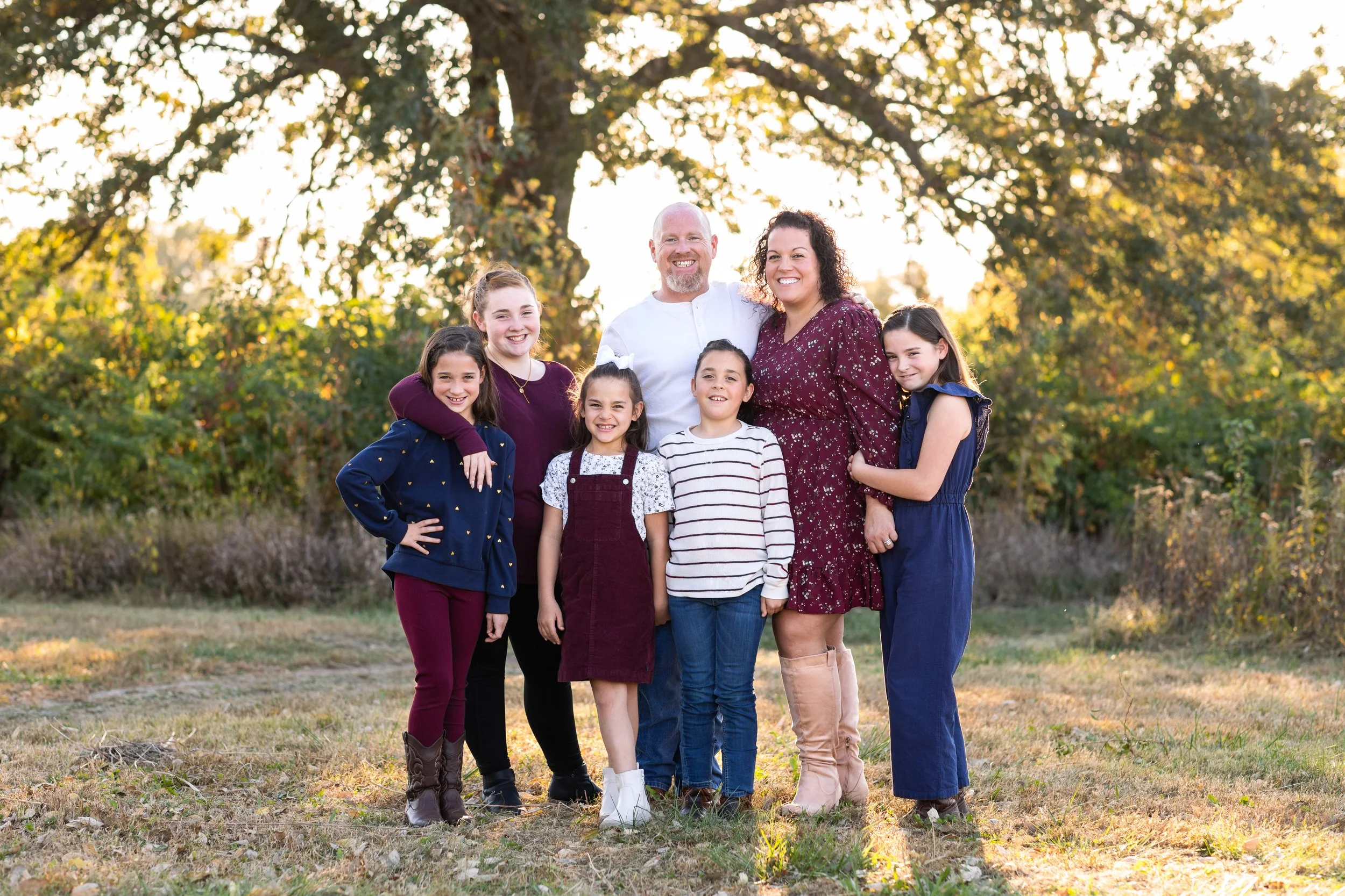 A smiling family of seven, including parents and five children, standing outdoors in a park during autumn, with trees and sunlight in the background.