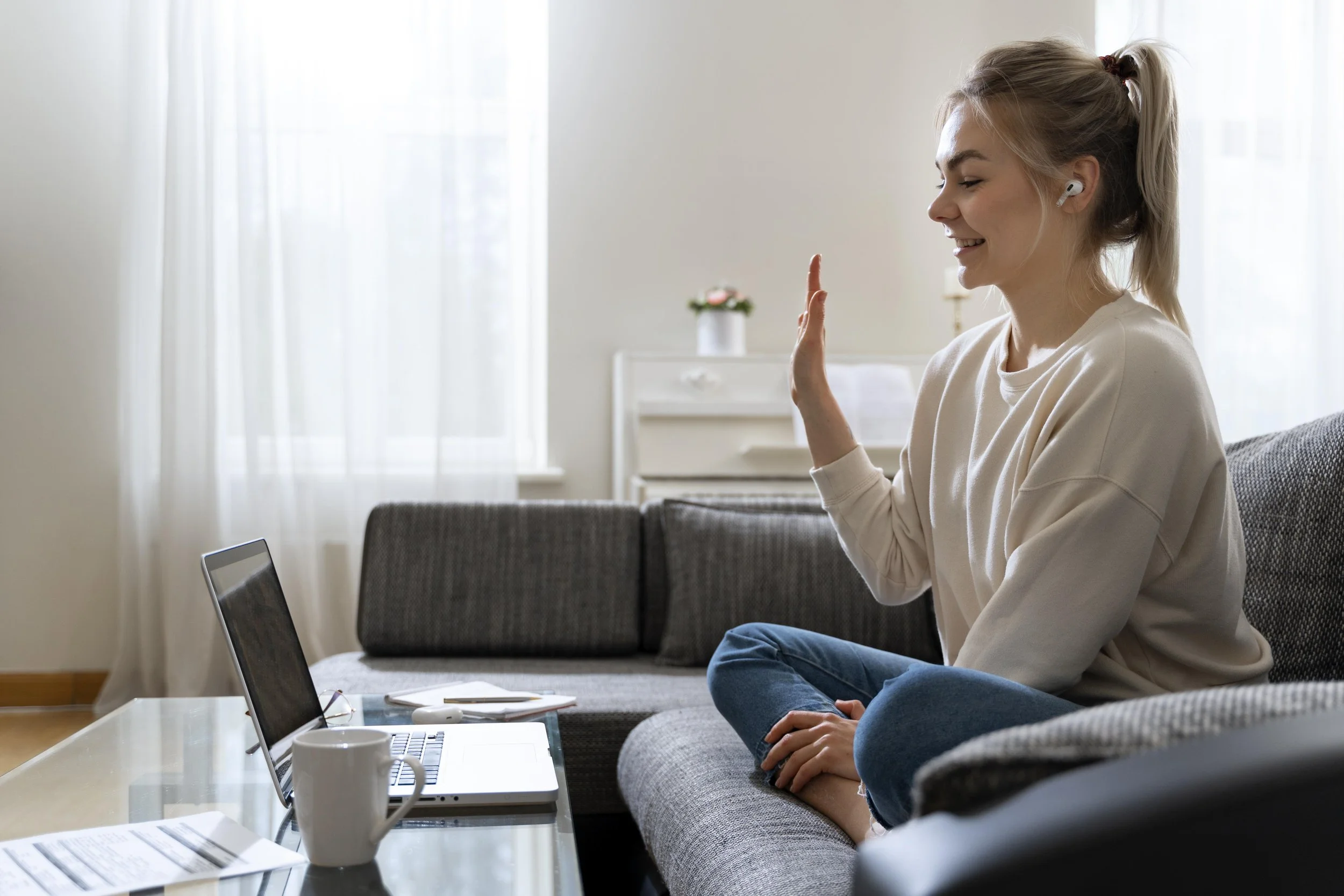 A woman with long blonde hair in a ponytail, wearing a white sweatshirt and earbuds, sitting on a gray sofa in a bright room with white curtains, waving at her laptop screen.