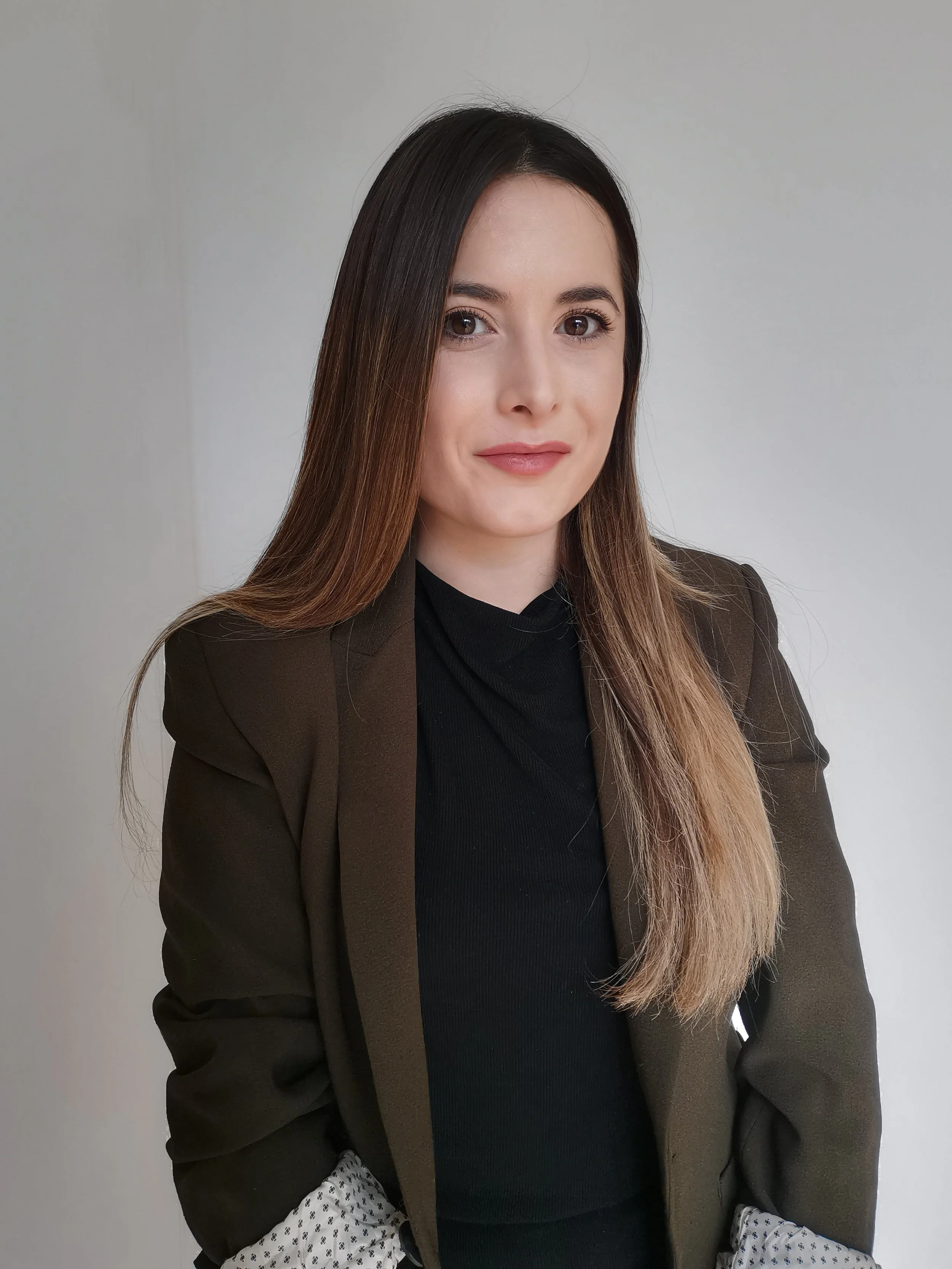 A woman with long brown hair, wearing a black turtleneck and a dark blazer, sitting against a plain light-colored wall.