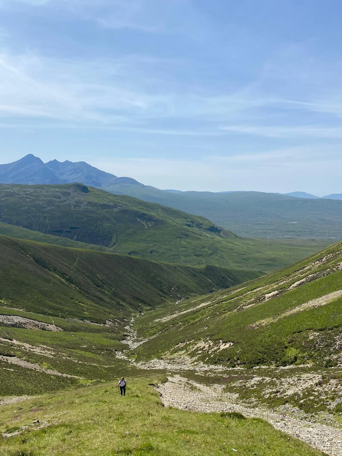 A person hiking in a lush green valley surrounded by rolling hills and mountains under a partly cloudy sky.