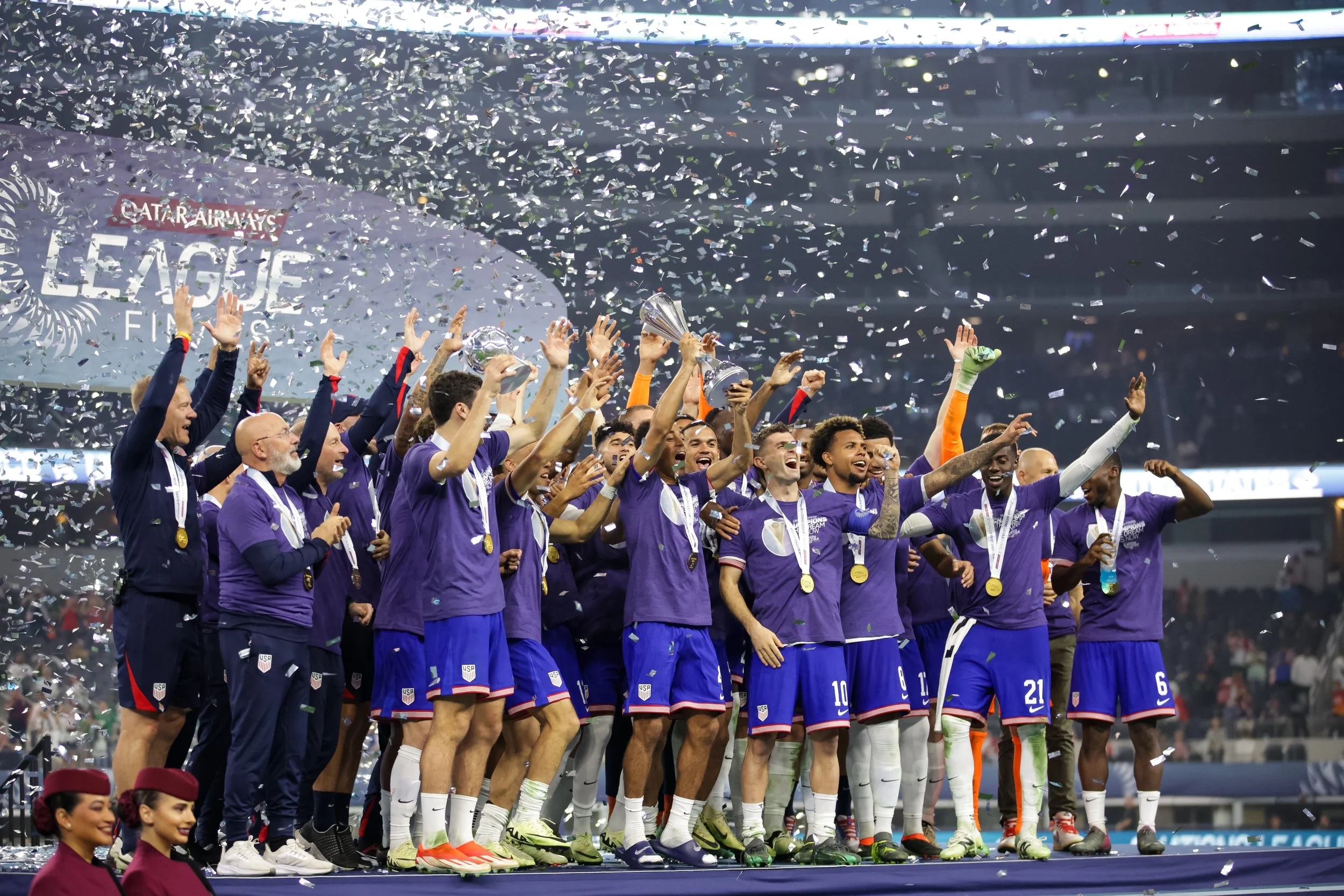 A group of soccer players and team staff celebrating on the field, holding trophies, with confetti falling around them.