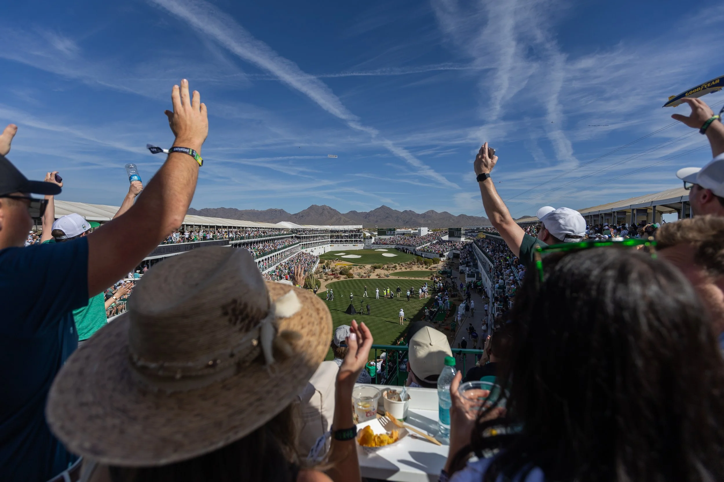 Crowd of spectators at a golf tournament, watching a hole on a sunny day with mountains and blue sky in the background.