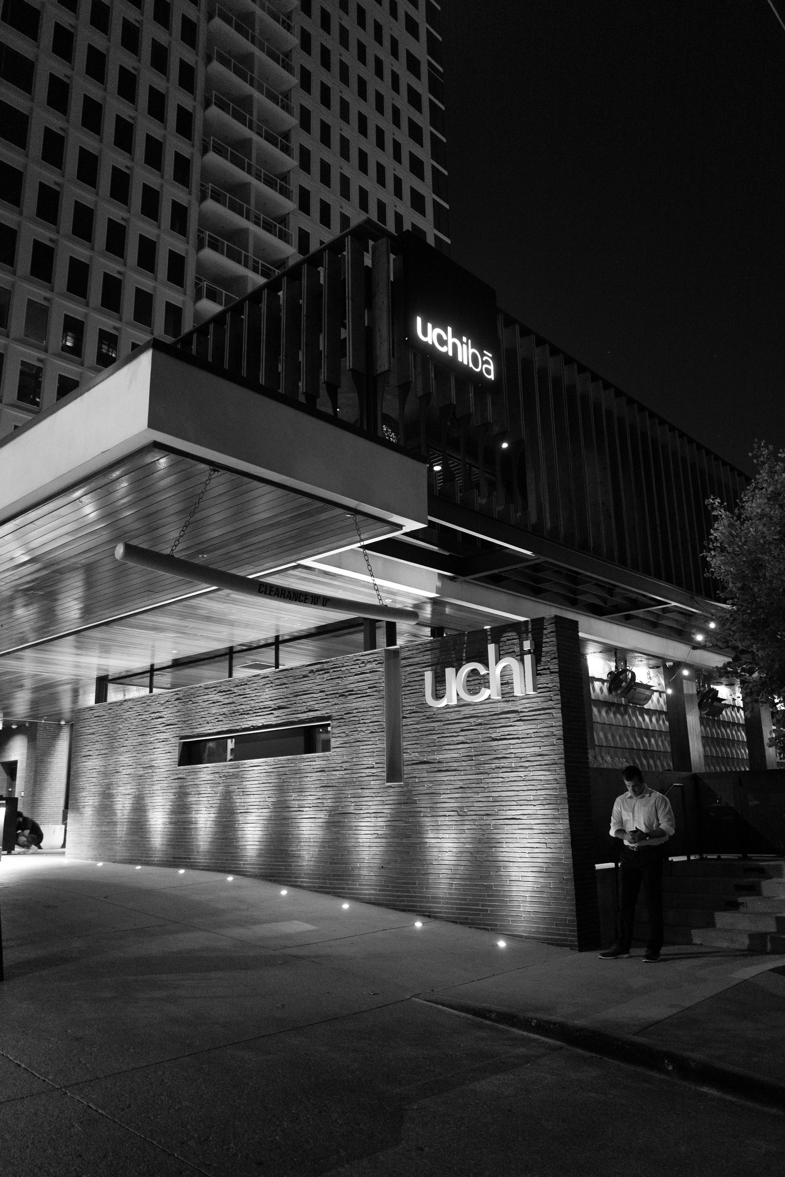 Night view of a modern building with illuminated signs reading 'Uchiôa' and 'uchi'. A man in a light-colored shirt is standing outside the building looking at his phone and another person is sitting on the ground nearby. The scene is in black and whi
