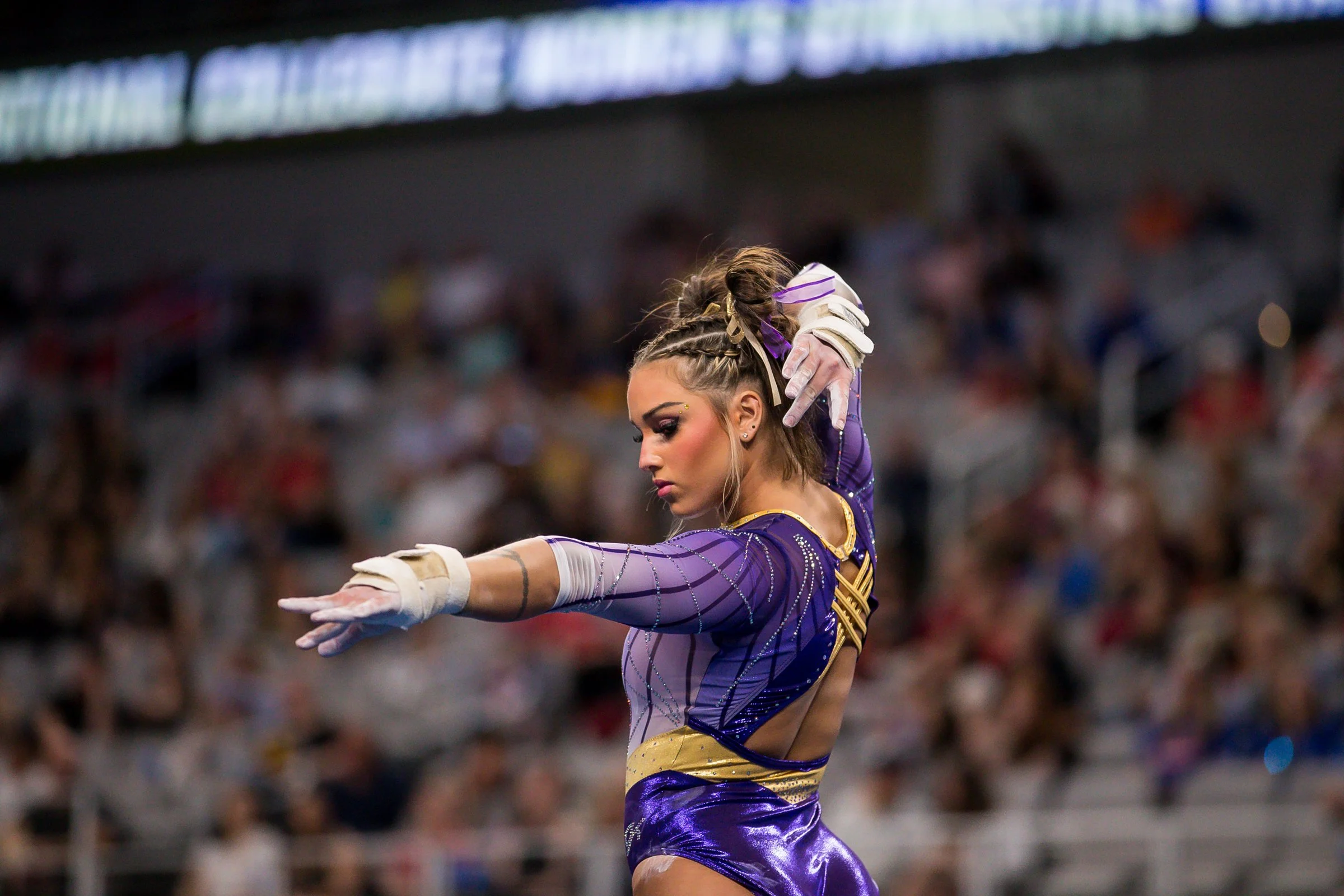 Female gymnast in purple and gold leotard performing a balancing routine in a gymnasium filled with spectators.