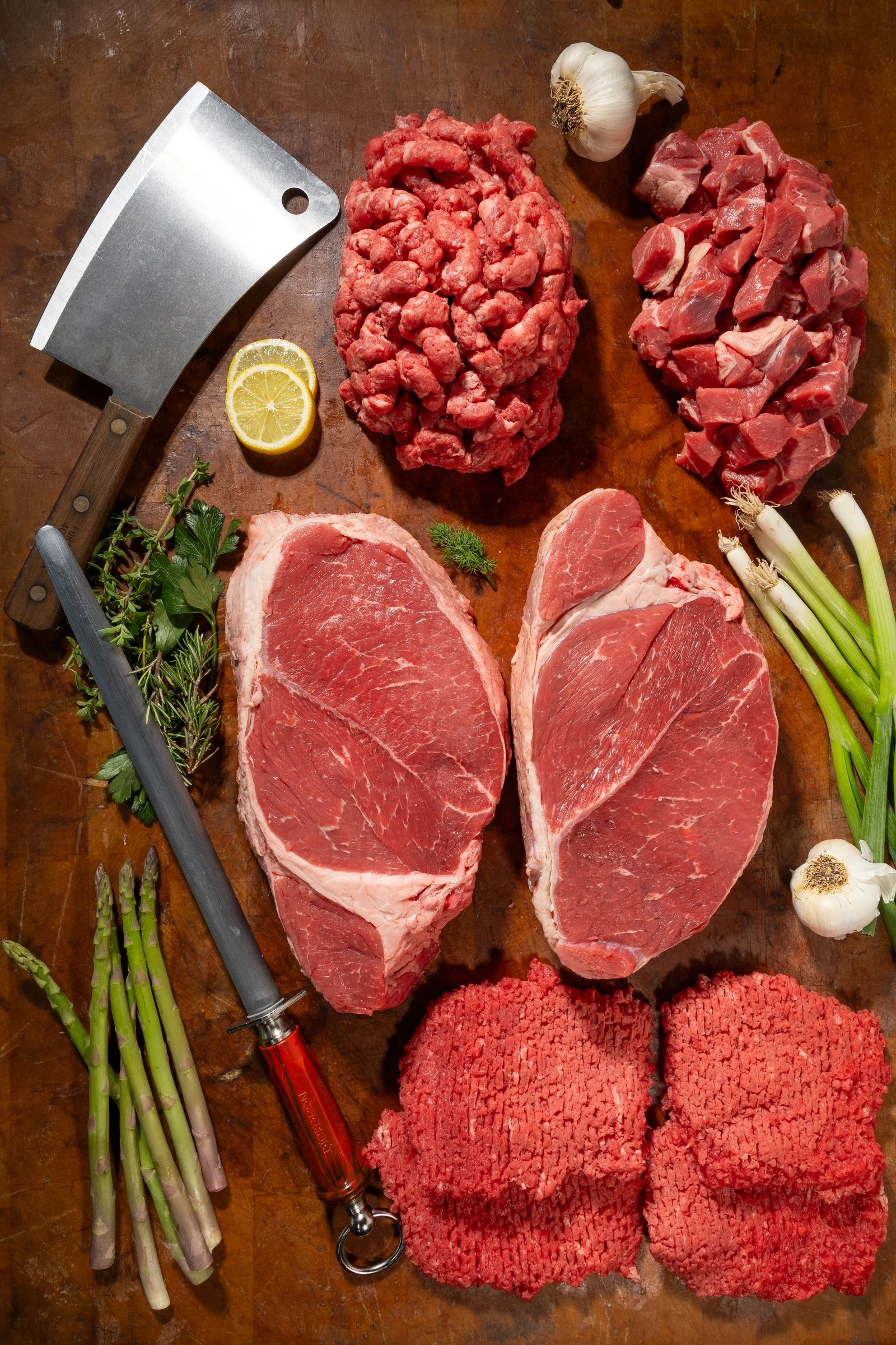 An overhead view of raw ingredients on a wooden cutting board, including three cuts of beef, chopped beef, garlic, lemon slices, green onions, herbs, and various kitchen knives.