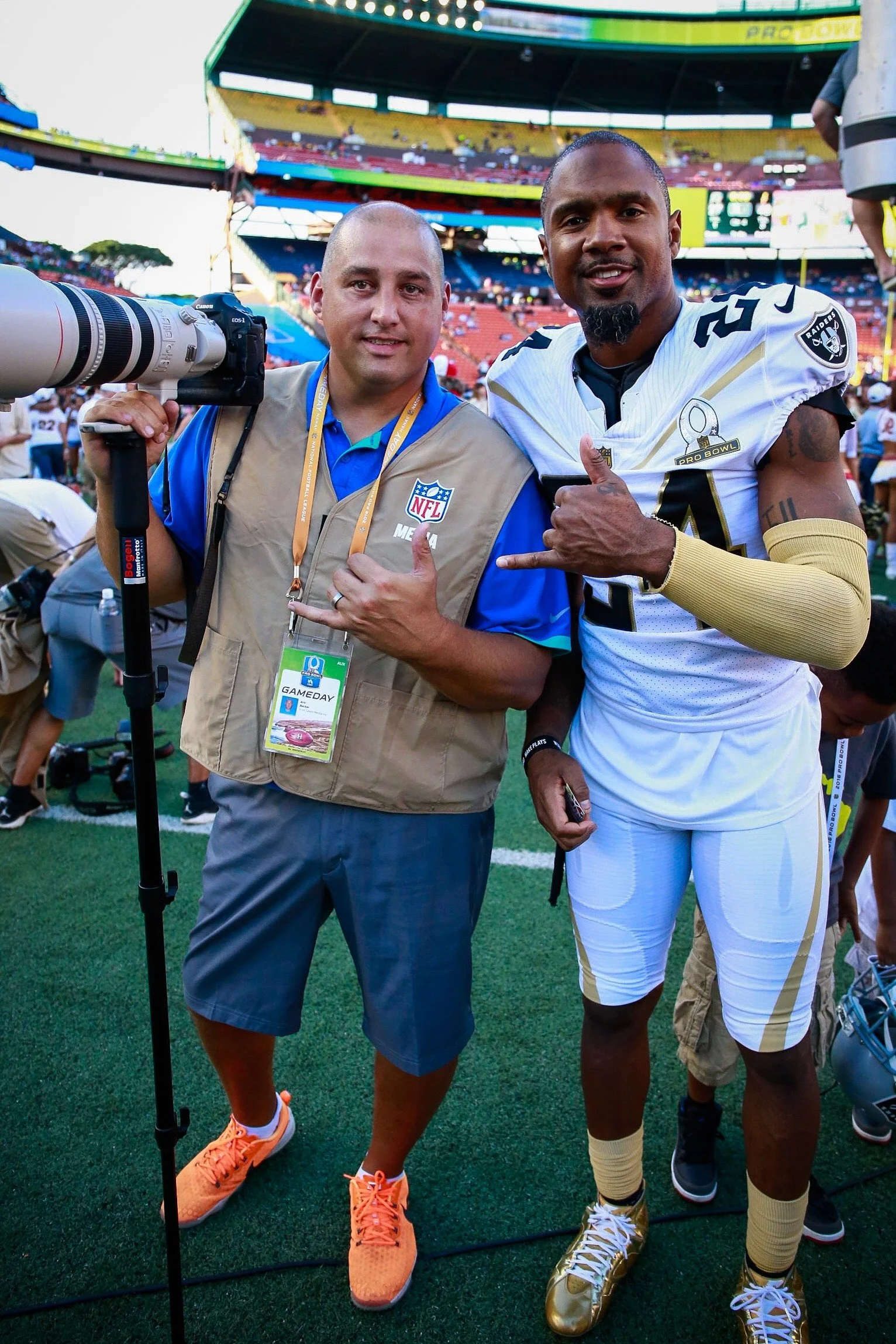 Two men on a football field posing for a photo, one is a photographer with a camera and the other is a football player in a black and white uniform with a Raiders logo.