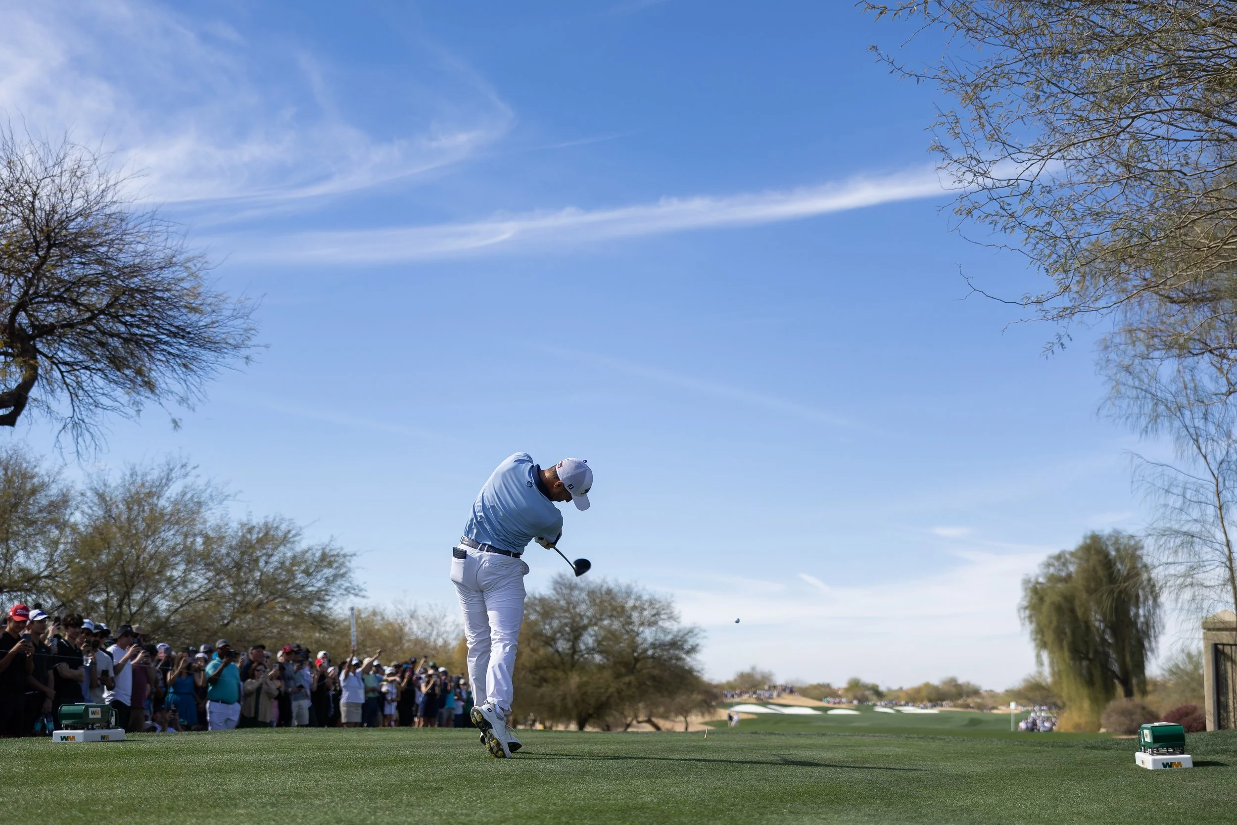 A golfer in a white shirt and white cap teeing off on a golf course with a crowd of spectators watching. The sky is clear with some clouds, and there are trees on the edges of the course.