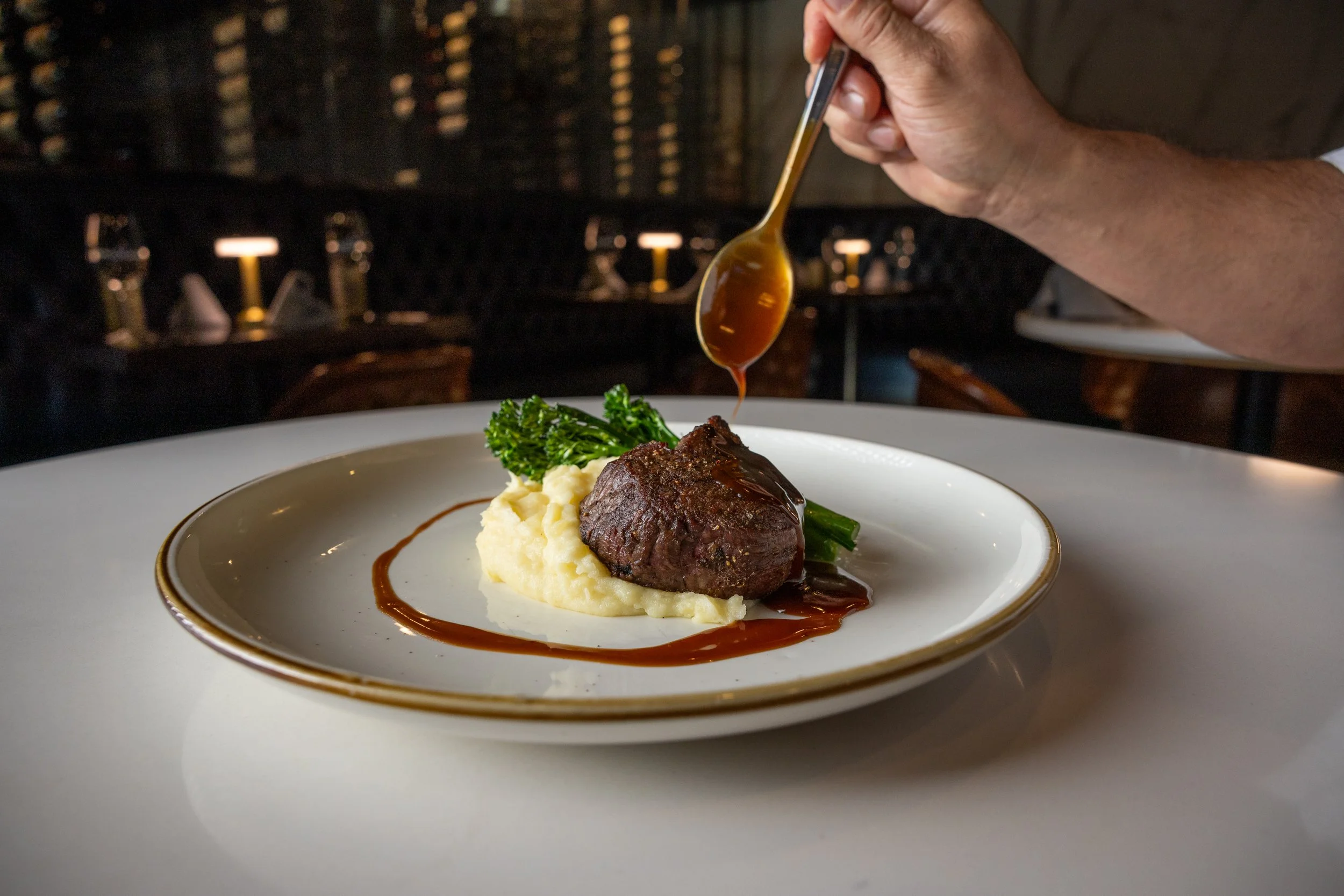 A plate with a serving of mashed potatoes topped with a piece of beef, garnished with green broccoli, and drizzled with brown gravy, with a hand pouring gravy from a spoon above the plate.
