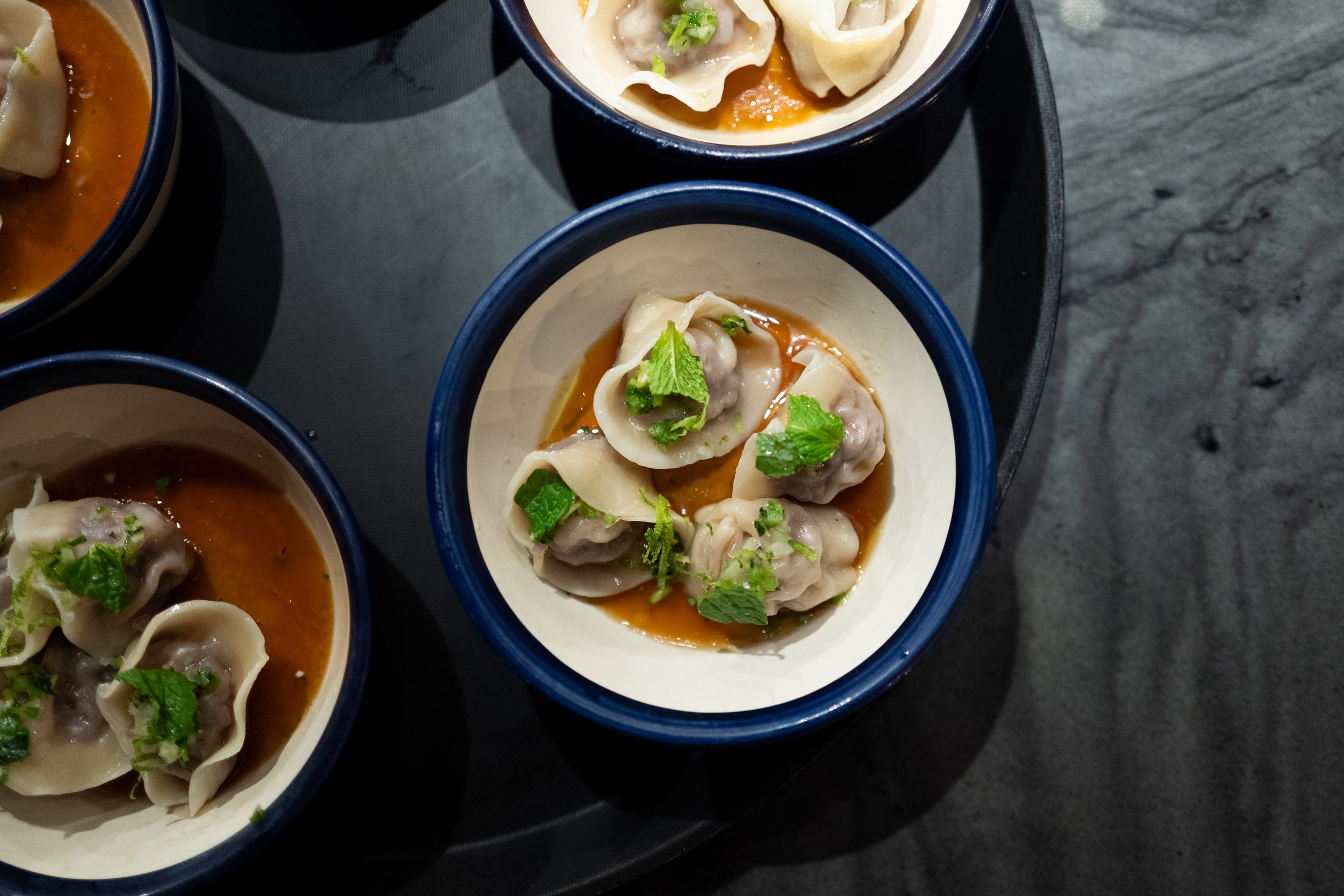 Close-up of steamed dumplings in white bowls with blue rims, garnished with chopped green herbs and sitting in a brown sauce on a black tray, placed on a dark gray surface.