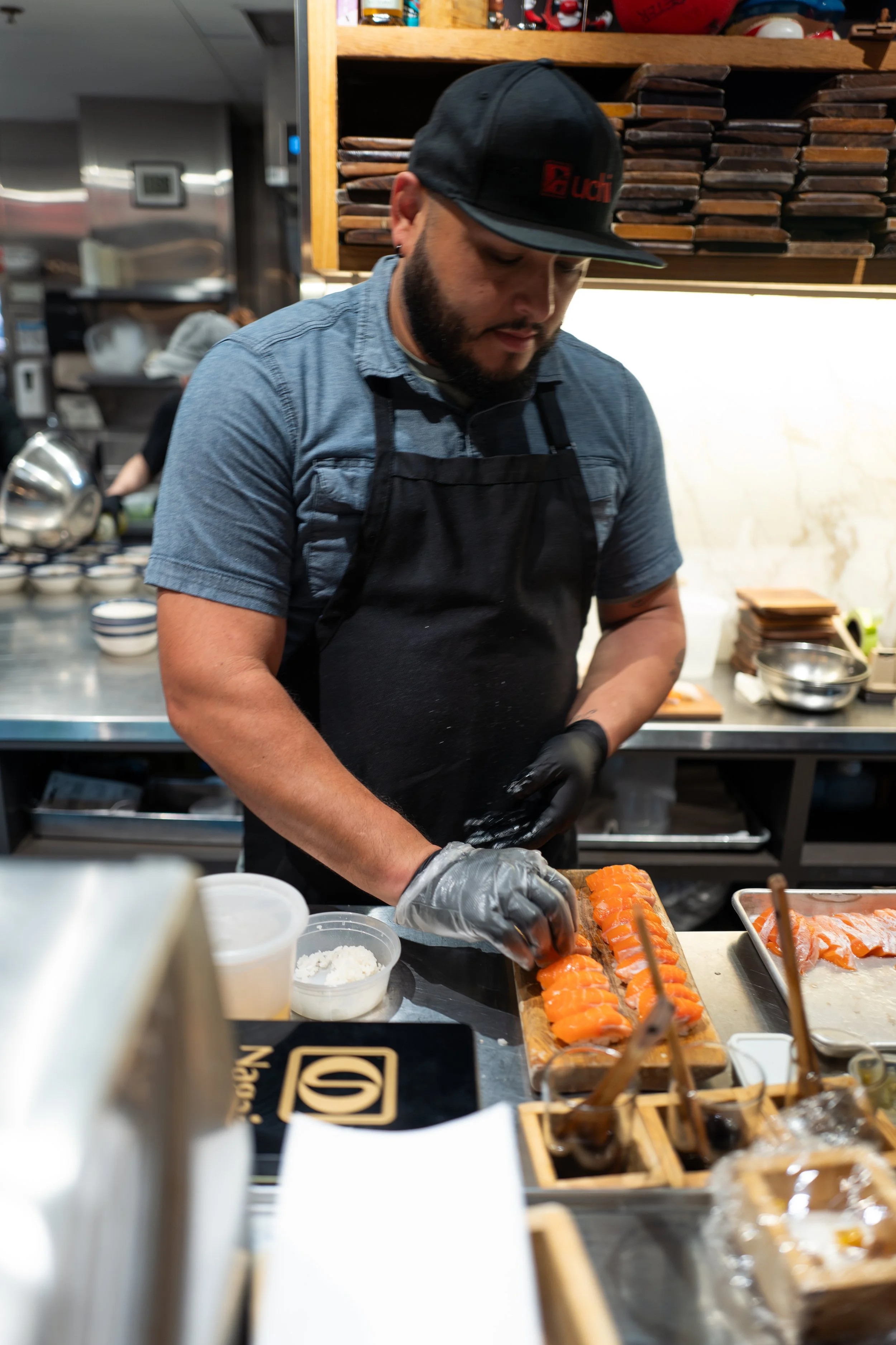 A man with a beard wearing a black cap, gray t-shirt, and black apron is preparing sushi in a kitchen. He is laying slices of salmon on a bamboo sushi rolling mat. The kitchen counter has bowls, containers of rice, and other sushi ingredients. In the