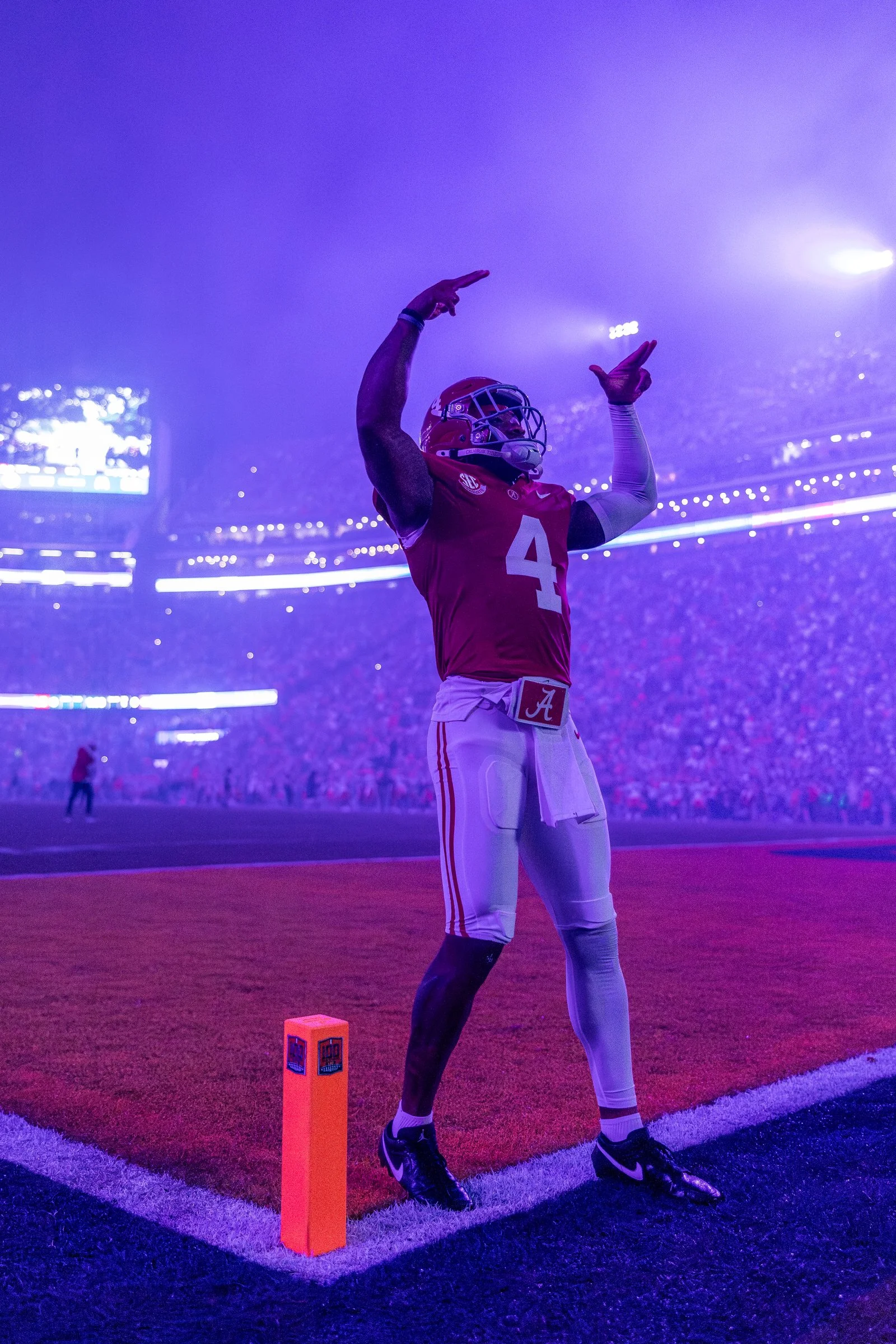 An American football player wearing a red jersey with the number 4 and white pants stands on the field near the end zone, preparing for a play under stadium lights with a crowd in the background.