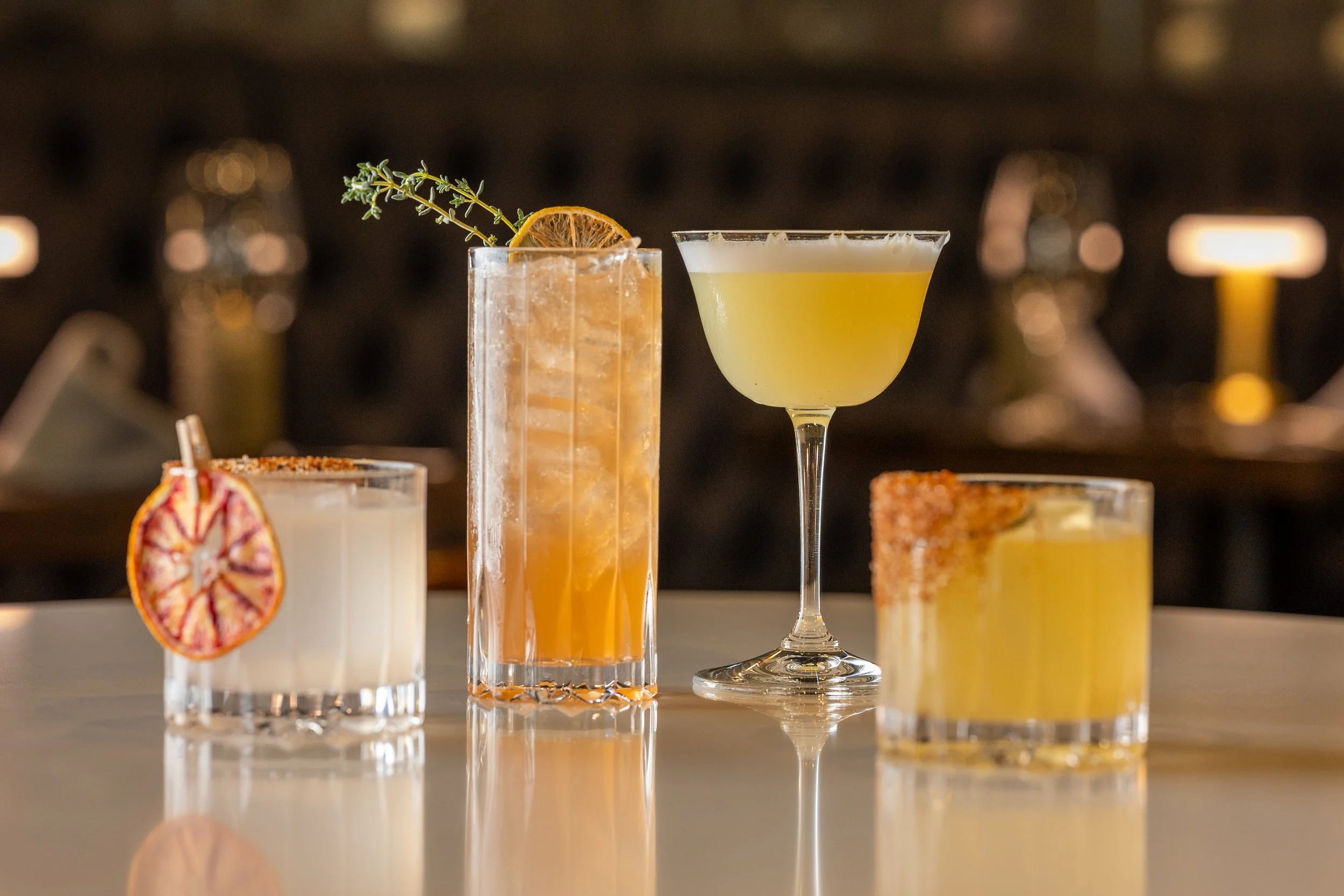 A variety of cocktails displayed on a bar counter, including a tall drink with an orange slice and thyme sprig, a yellow-green drink in a martini glass, and other beverages with garnishes.