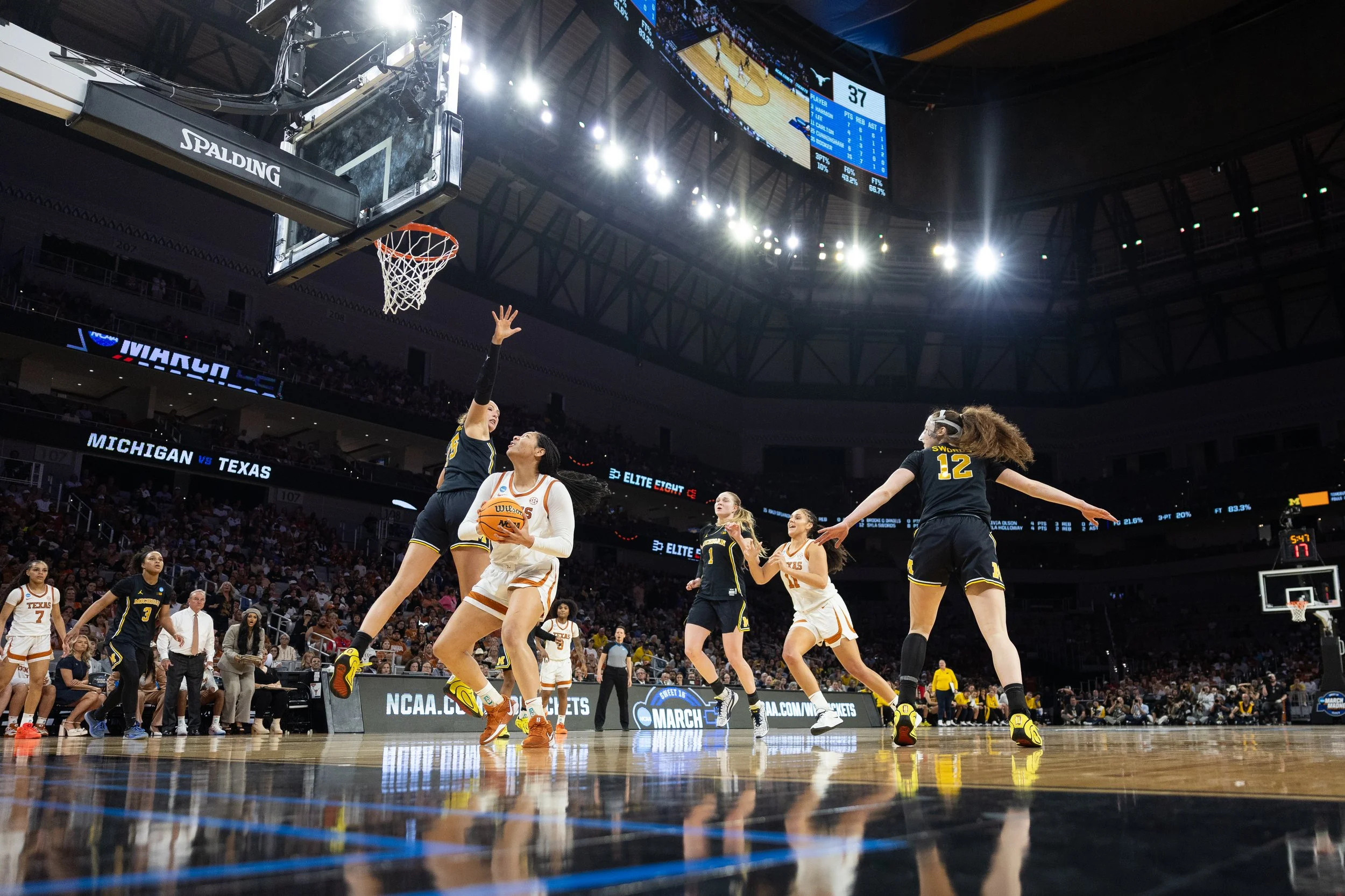 A women's basketball game in a stadium with players from two teams, Texas and Michigan, actively playing. One player from Michigan is holding the ball while others are reaching for a rebound or defending. The game is intense, and the background revea
