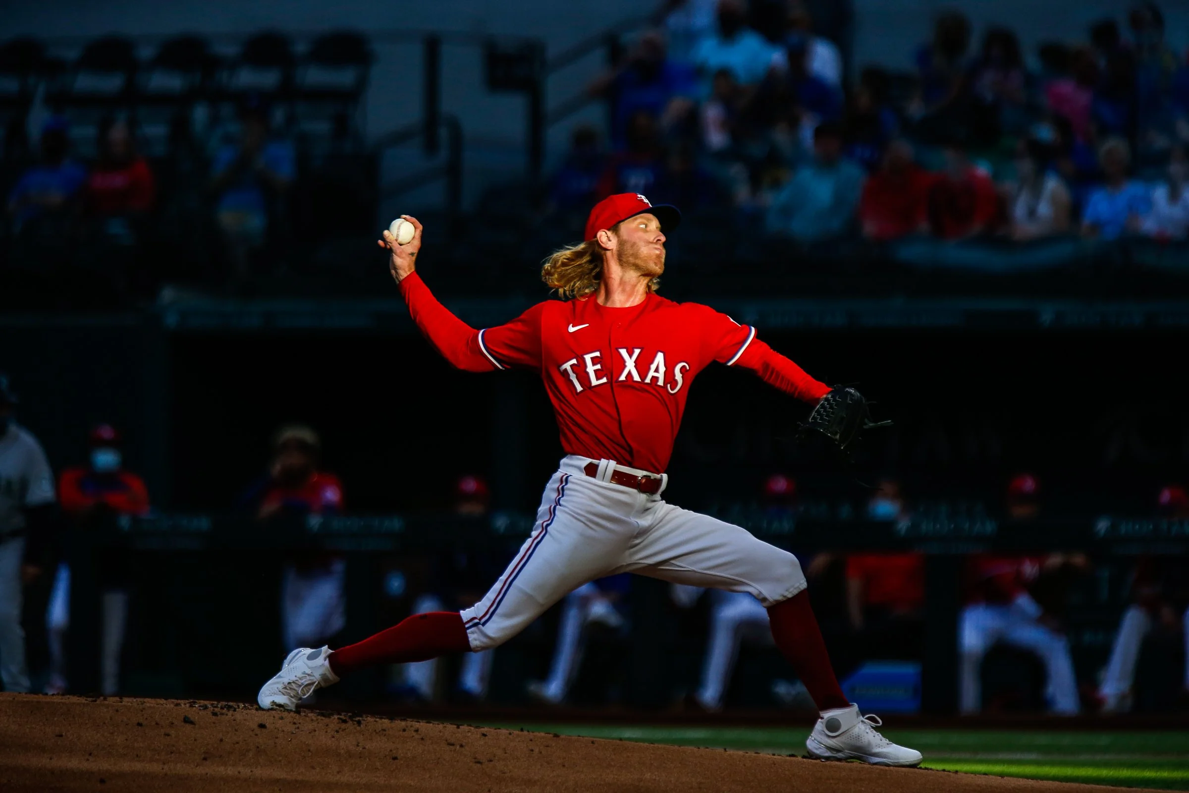 A baseball player in a red Texas Rangers uniform throws a pitch on the field during a game at night, with spectators in the stands in the background.