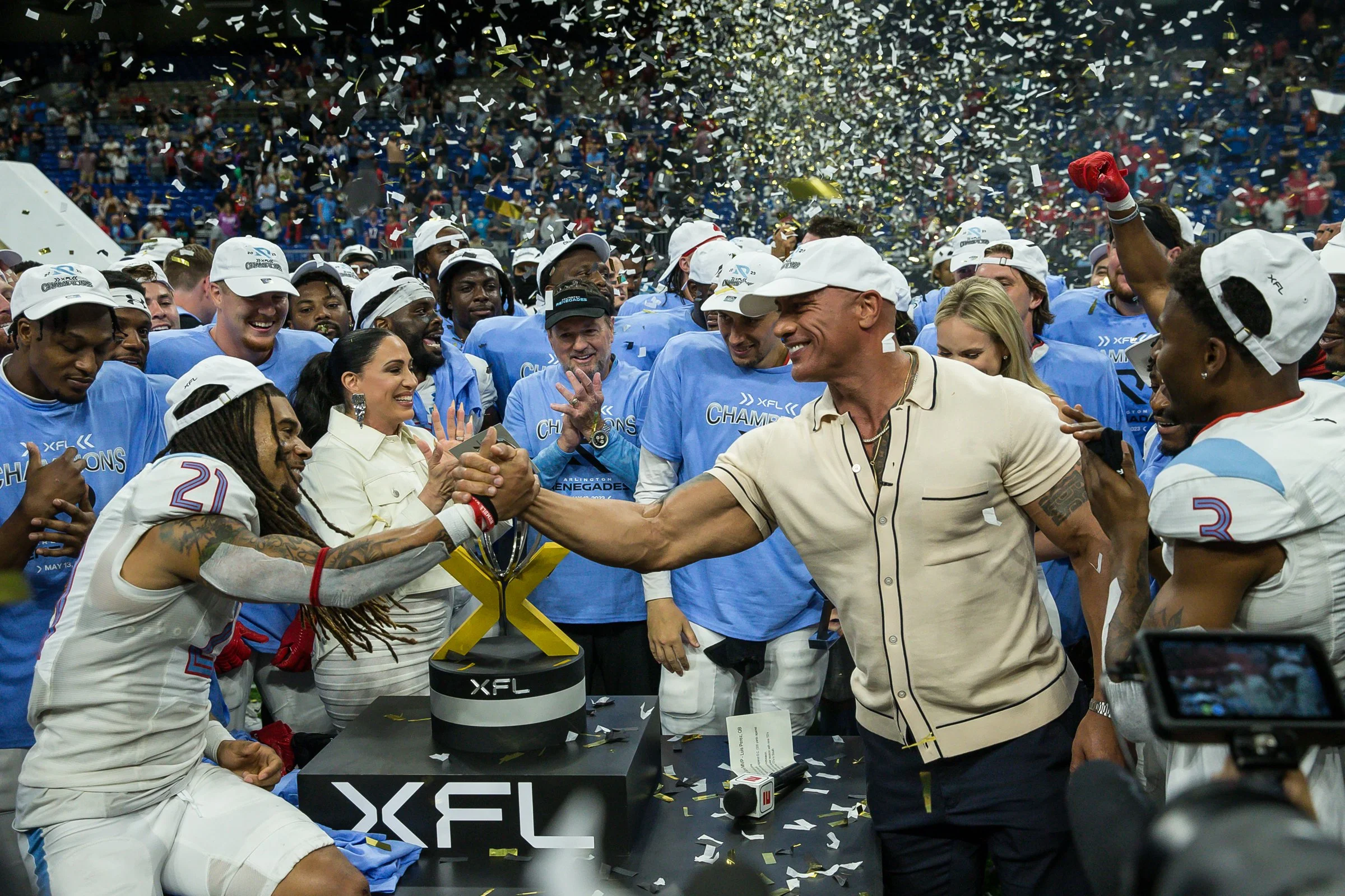 A group of people celebrating on a sports field, with confetti falling from above. Some are wearing blue shirts and white caps, and they are gathered around a table with a trophy. People are smiling and shaking hands in a joyful atmosphere.