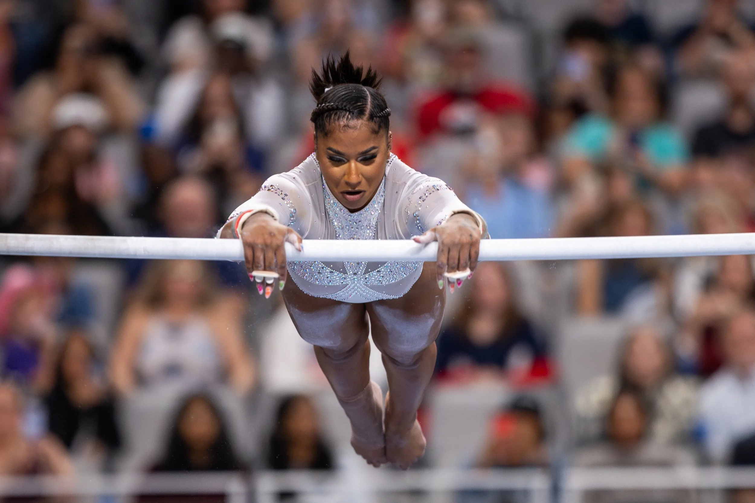 A female gymnast in a white, embellished leotard performs a routine on the balance beam, with a blurred audience in the background.