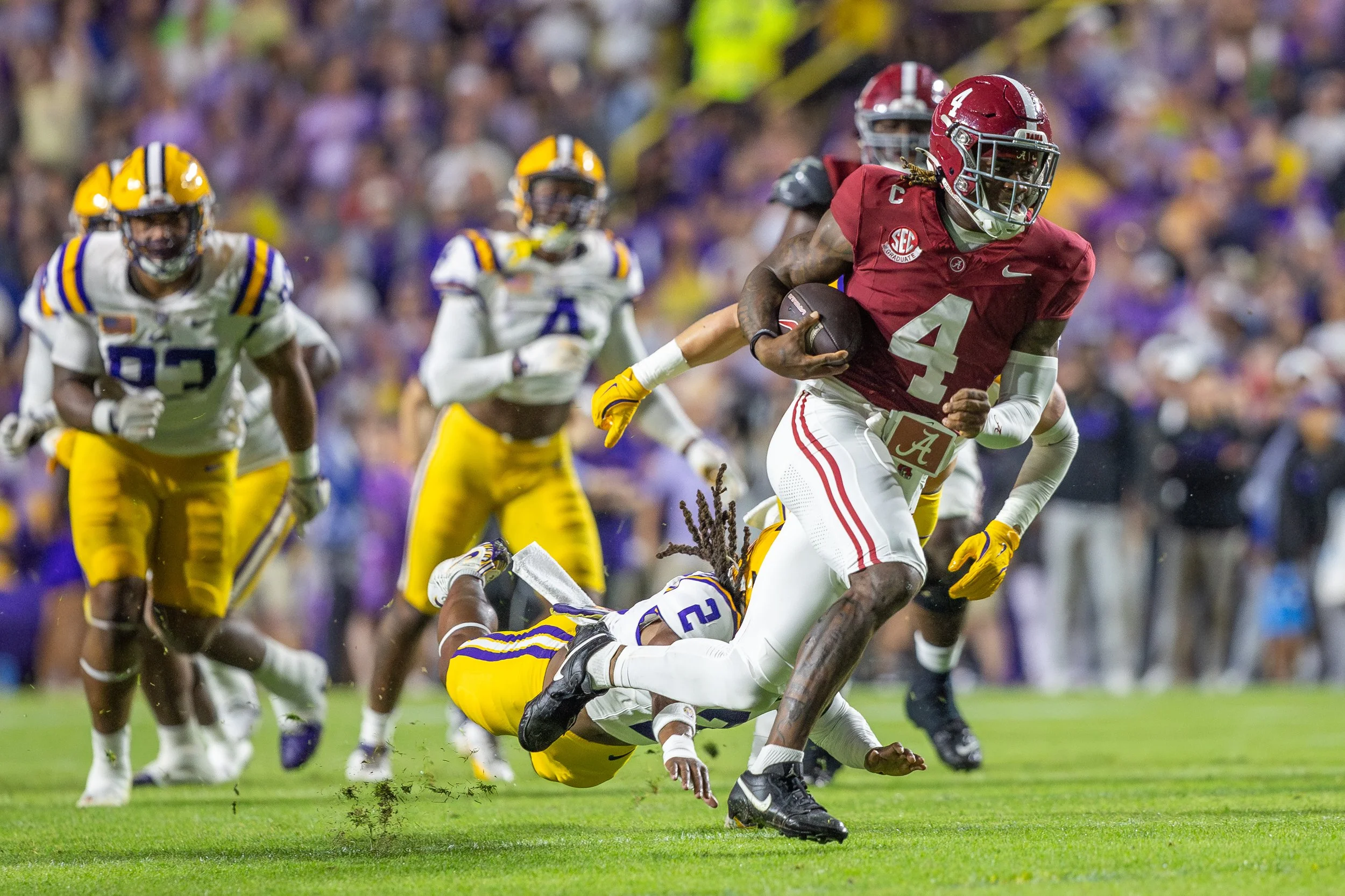 An Alabama football player wearing a red jersey with number 4 running with the ball while a player from LSU in a yellow and purple uniform falls to the ground during a game, with other players and spectators in the background.