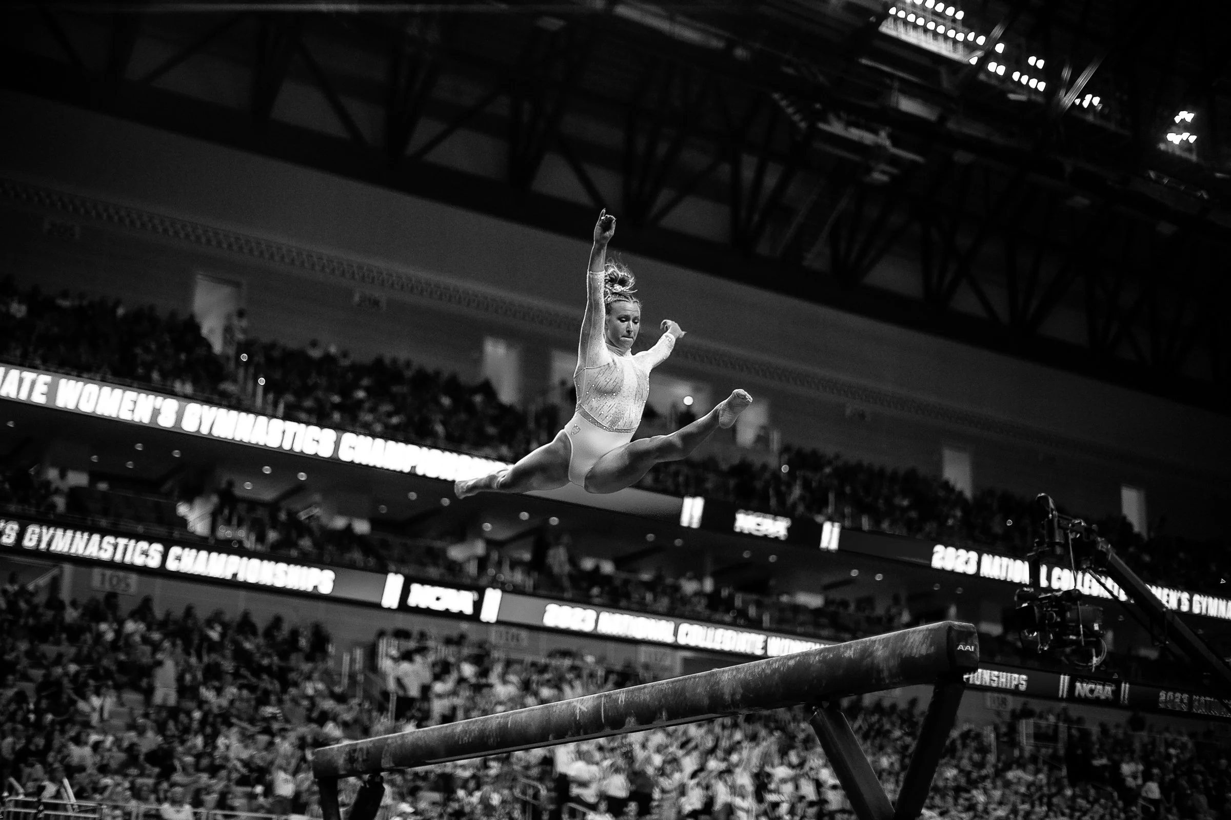 A female gymnast mid-air performing on the balance beam during a gymnastics competition in a large stadium filled with spectators.