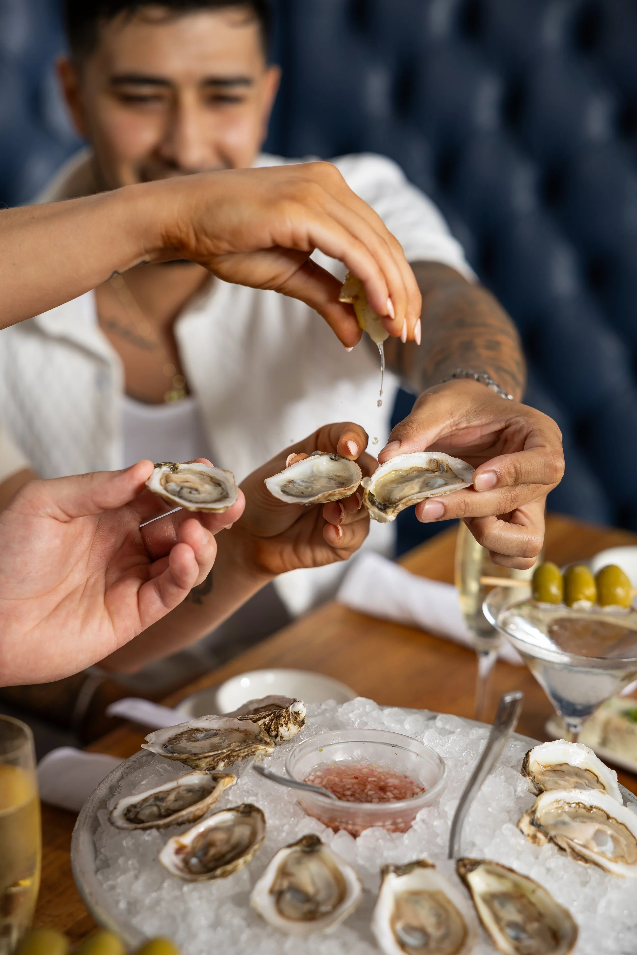 A person is holding an oyster in one hand while squeezing lemon over it with the other hand. The oyster is on a tray filled with ice and other oysters. There are also small bowls of sauce and a glass with a drink in the background.