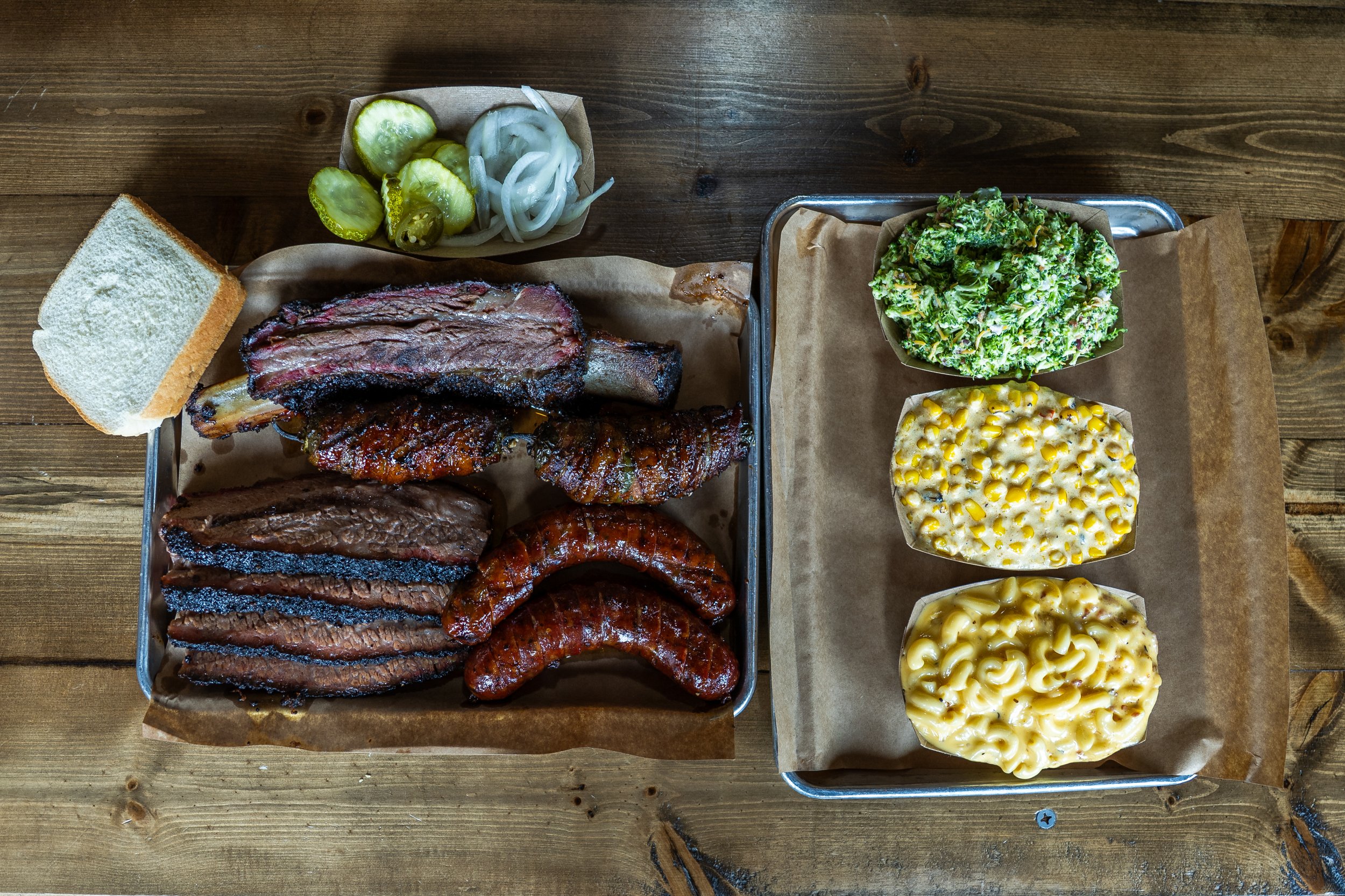 A top view of a wooden table with a metal tray of assorted barbecue ribs and sliced bread, alongside a small basket of pickles and onions, and a basket of three bowls of macaroni and cheese, broccoli salad, and coleslaw.