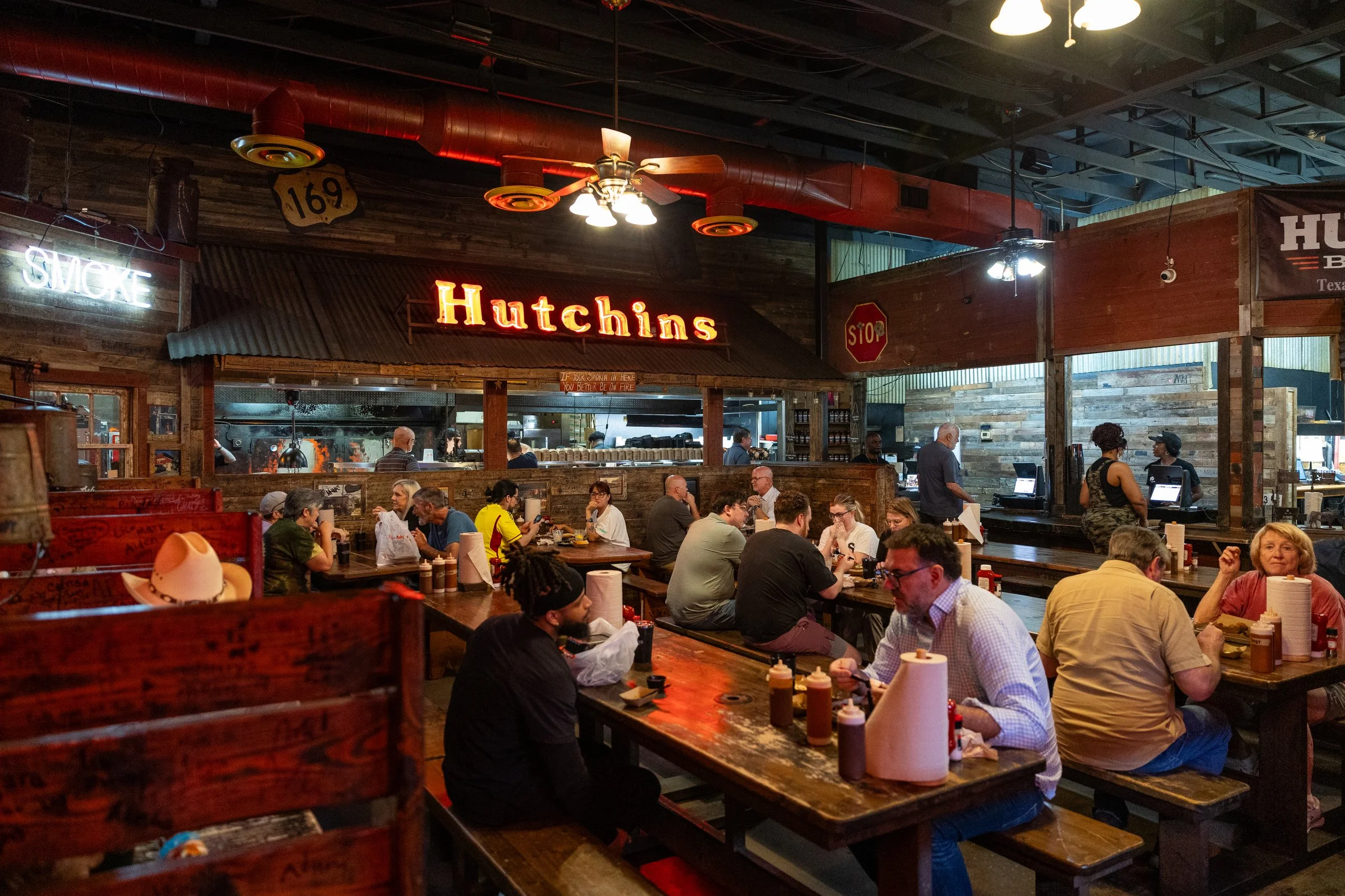 The interior of a busy restaurant with wooden decor, a lively crowd of diners, and neon signs including a prominent 'Hutchins' sign. People are seated at long tables, some wearing cowboy hats, and staff are seen working behind the counter.