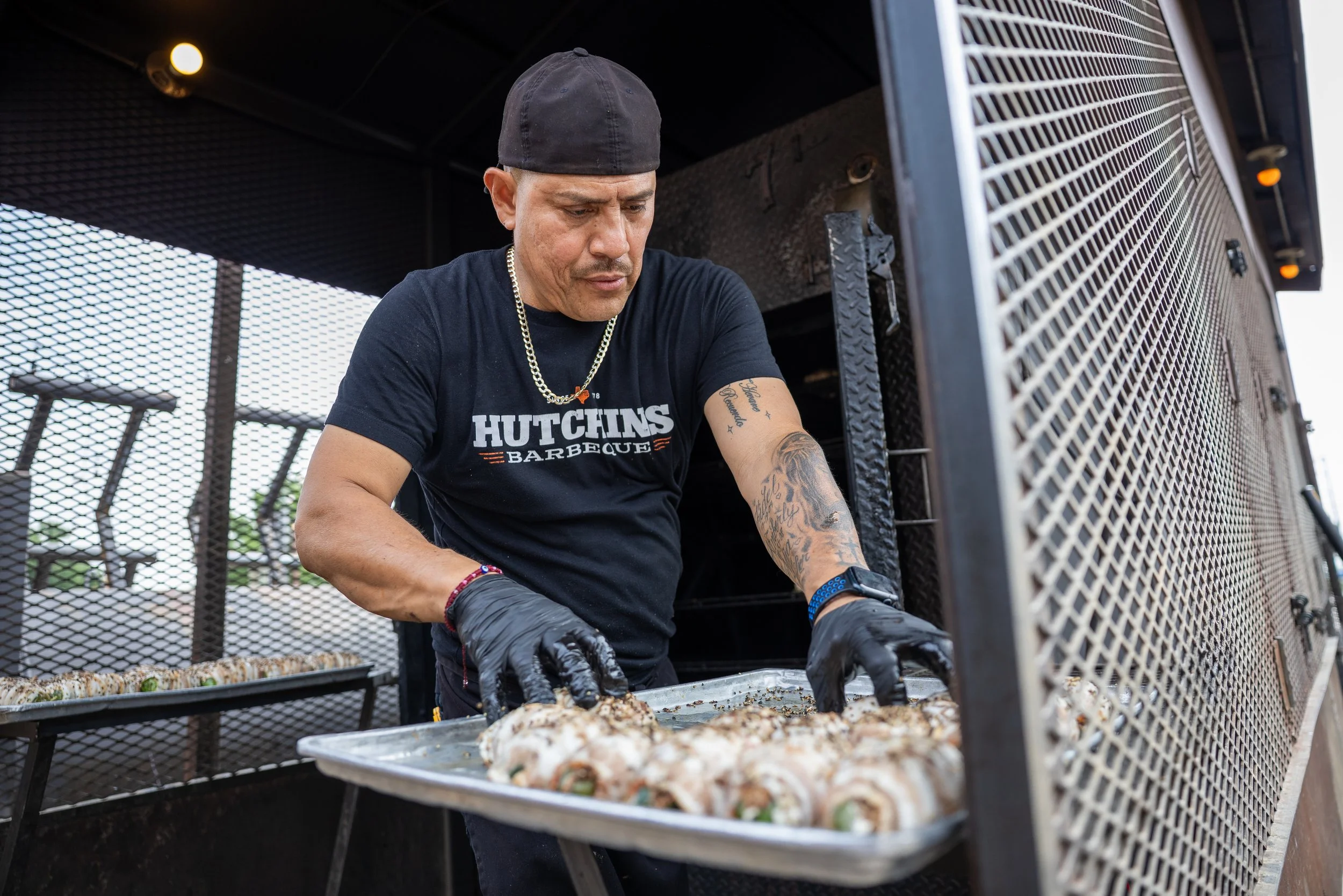 A man wearing a black hat, black t-shirt with 'HUTCHINS BARBEQUE' written on it, and gloves is preparing food on a grill outdoors. The grill has a metal exterior with a grated surface, and there are trays of food in front of him.