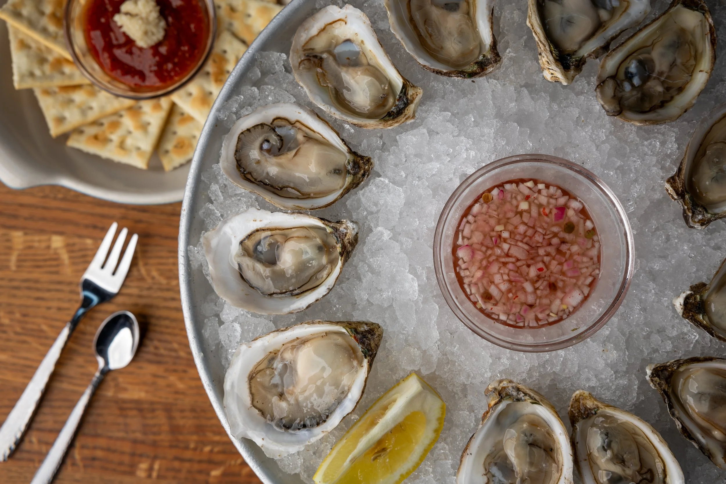 A seafood platter featuring fresh oysters on ice, lemon wedges, and cocktail sauce with diced onions and peppers, arranged on a metal tray. There are also crackers with a red dipping sauce and a small fork visible on a wooden surface.