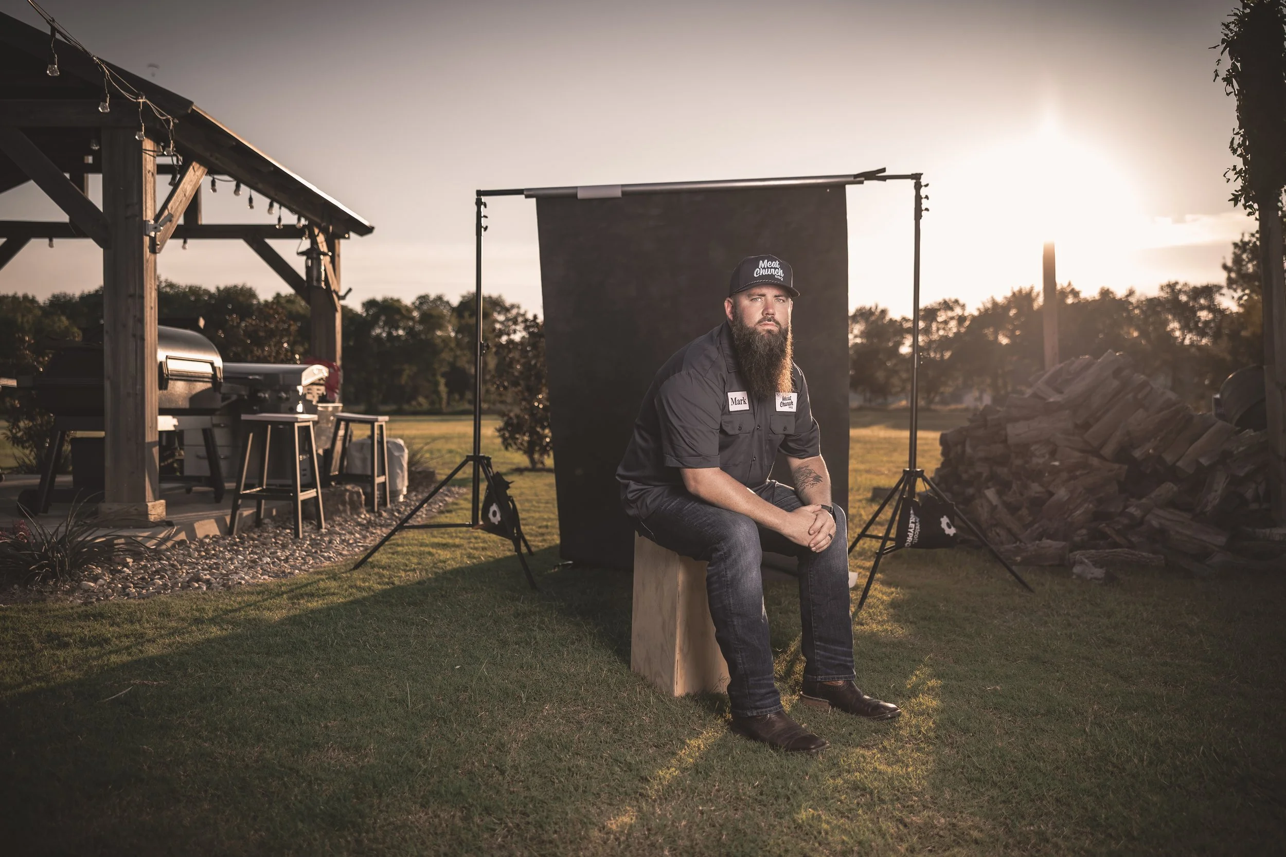 A man with a beard, wearing a black cap and black shirt, sits on a wooden stool outdoors during sunset. Behind him, photography equipment including a backdrop and lighting is set up. There is a pile of logs on his right, and trees in the background.