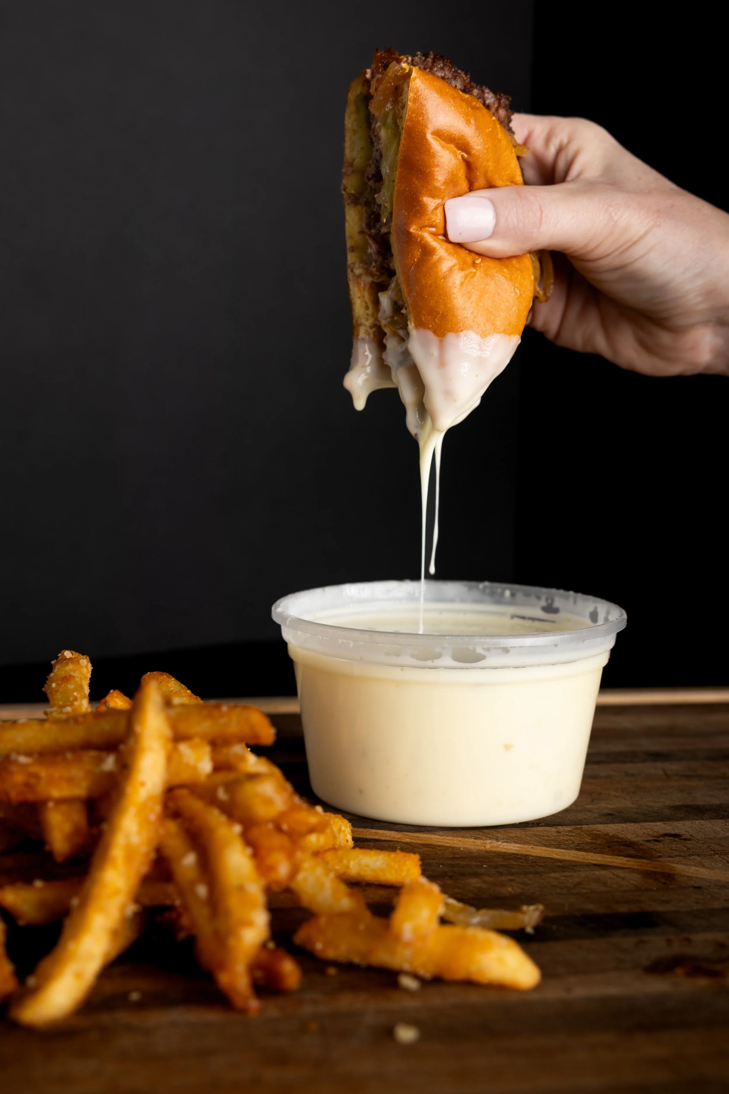 Hand holding a cheeseburger with melted cheese dripping, French fries with seasoning, and a cup of dipping sauce on a wooden surface against a black background.