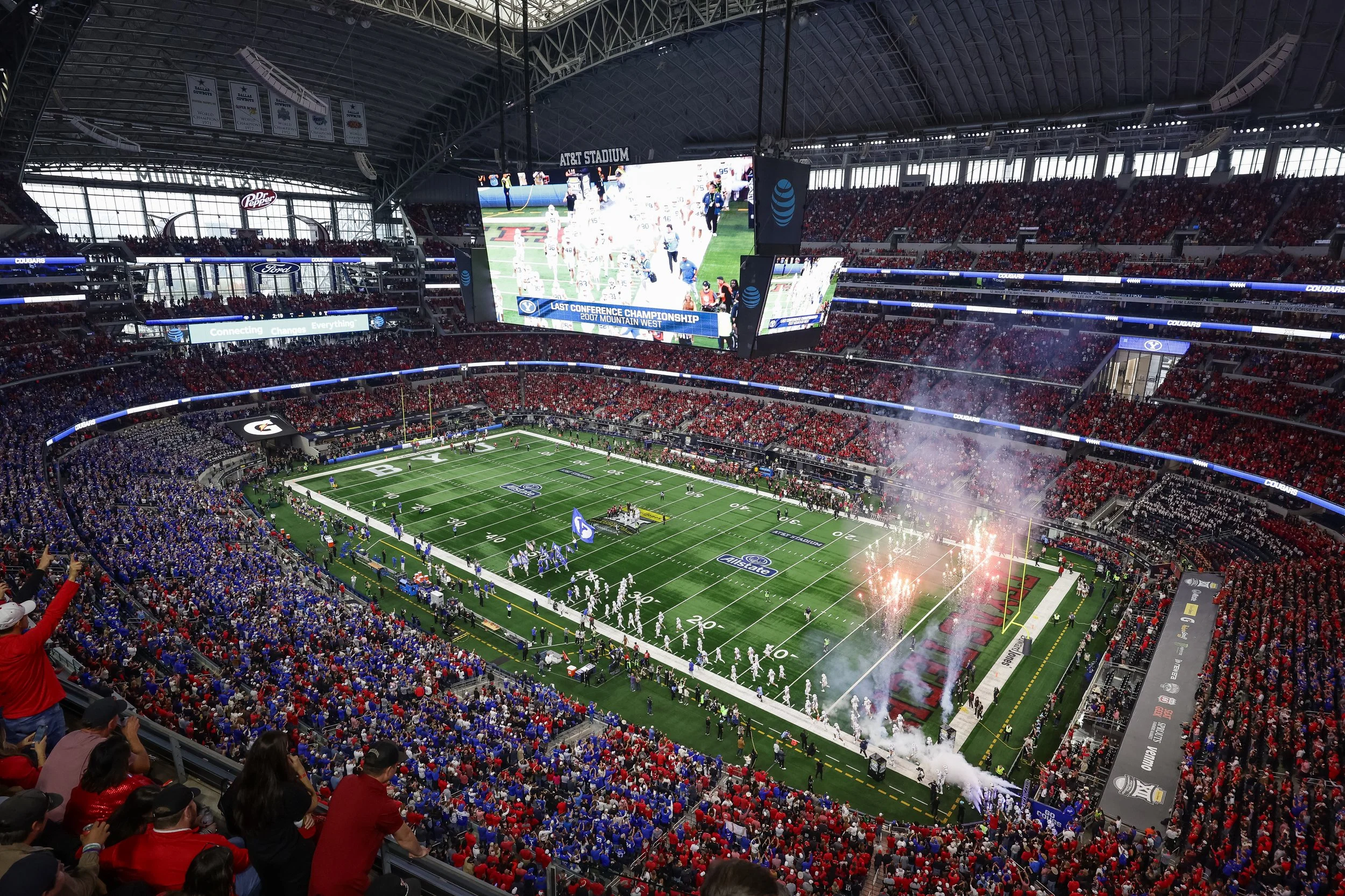 Interior view of AT&T Stadium during a football game, with players on the field, large video screens displaying the game, and thousands of spectators dressed in team colors seated in the stands.