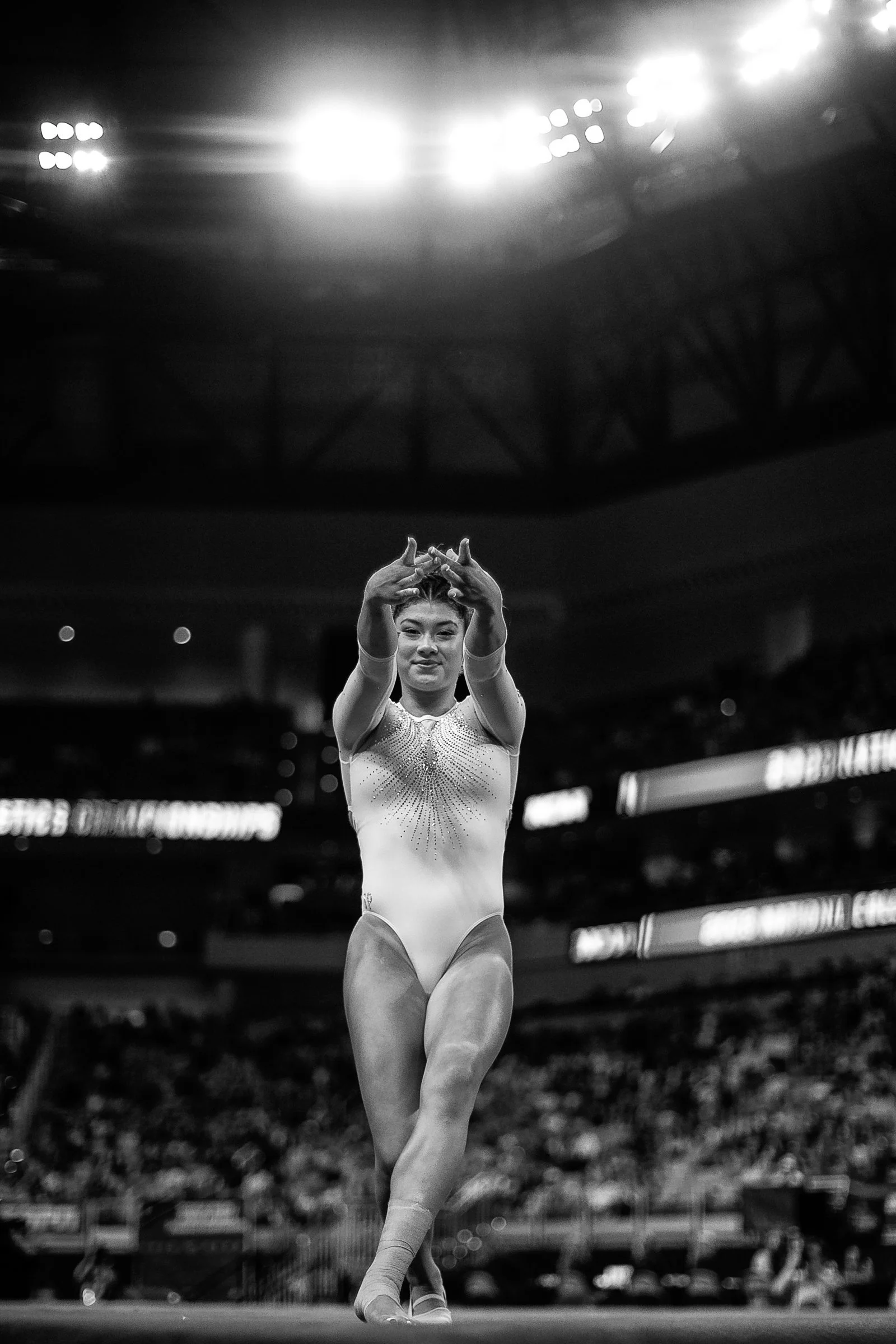 Black and white photo of a female gymnast performing a routine on the balance beam in an indoor arena, with bright stadium lights overhead.
